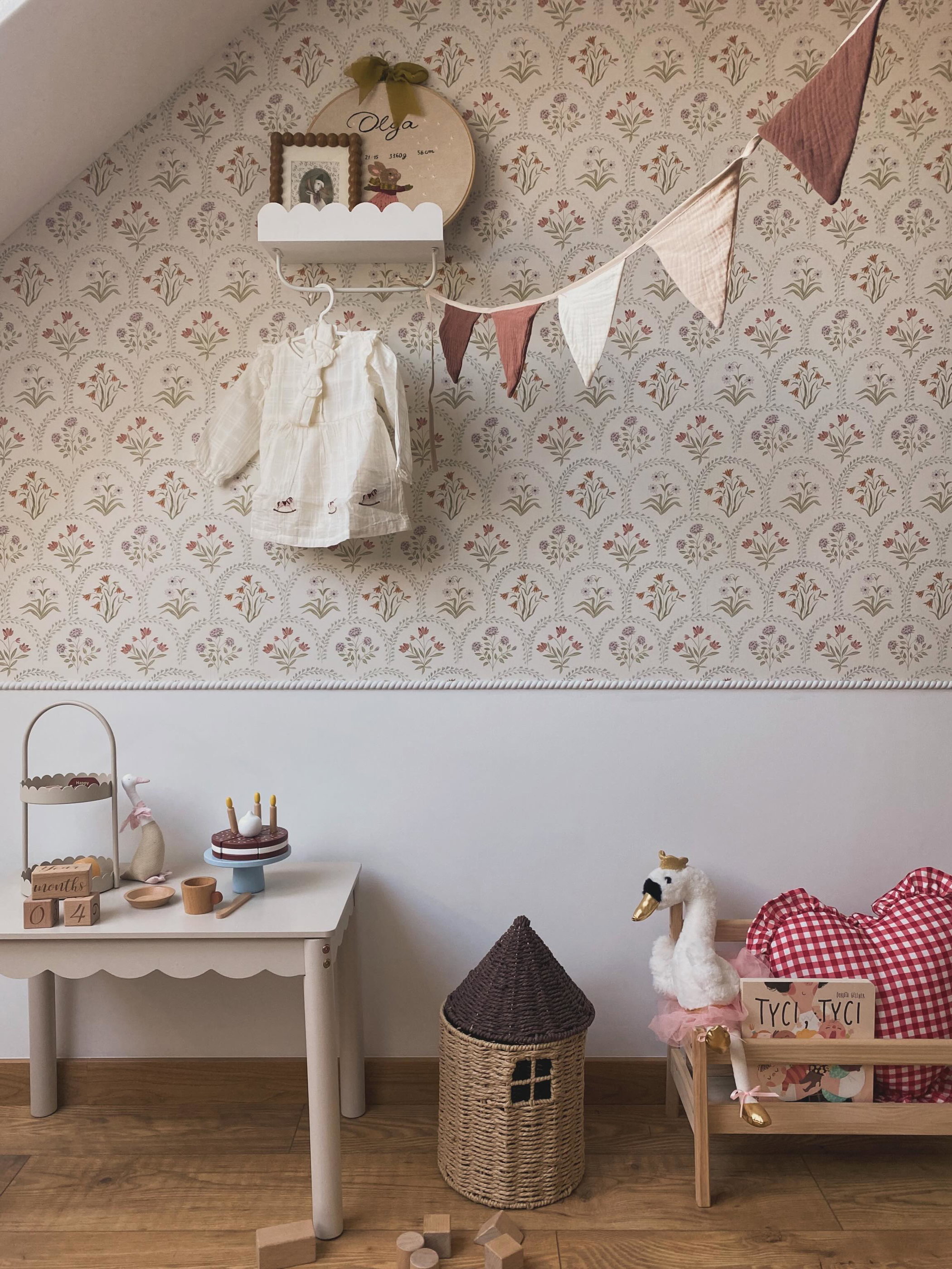 A child's bedroom decorated with floral wallpaper, featuring a small table with a cake, a swan plushie on a bed, and a woven basket.