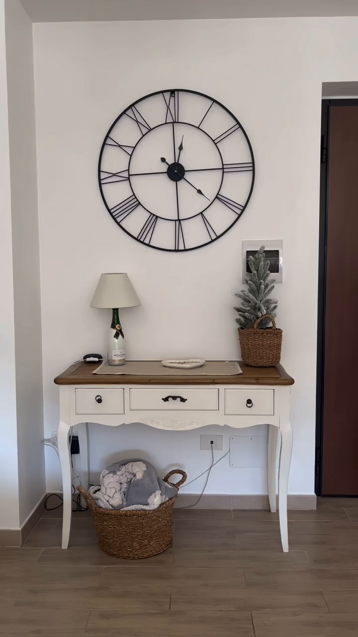 Entryway table with decor. Large clock hangs above a white table with lamp, tree, and basket.