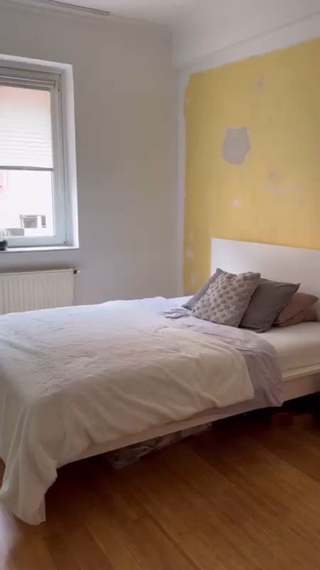 Bedroom with white bed, gray pillows, and yellow accent wall with peeling paint.