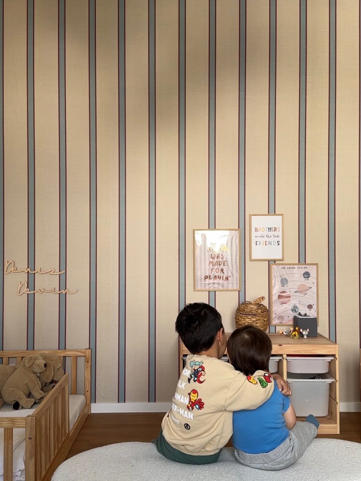 Dos niños, vistos desde atrás, se sientan en una alfombra blanca con los brazos alrededor del otro. Miran una pared a rayas beige y gris con arte enmarcado y una cama de madera.