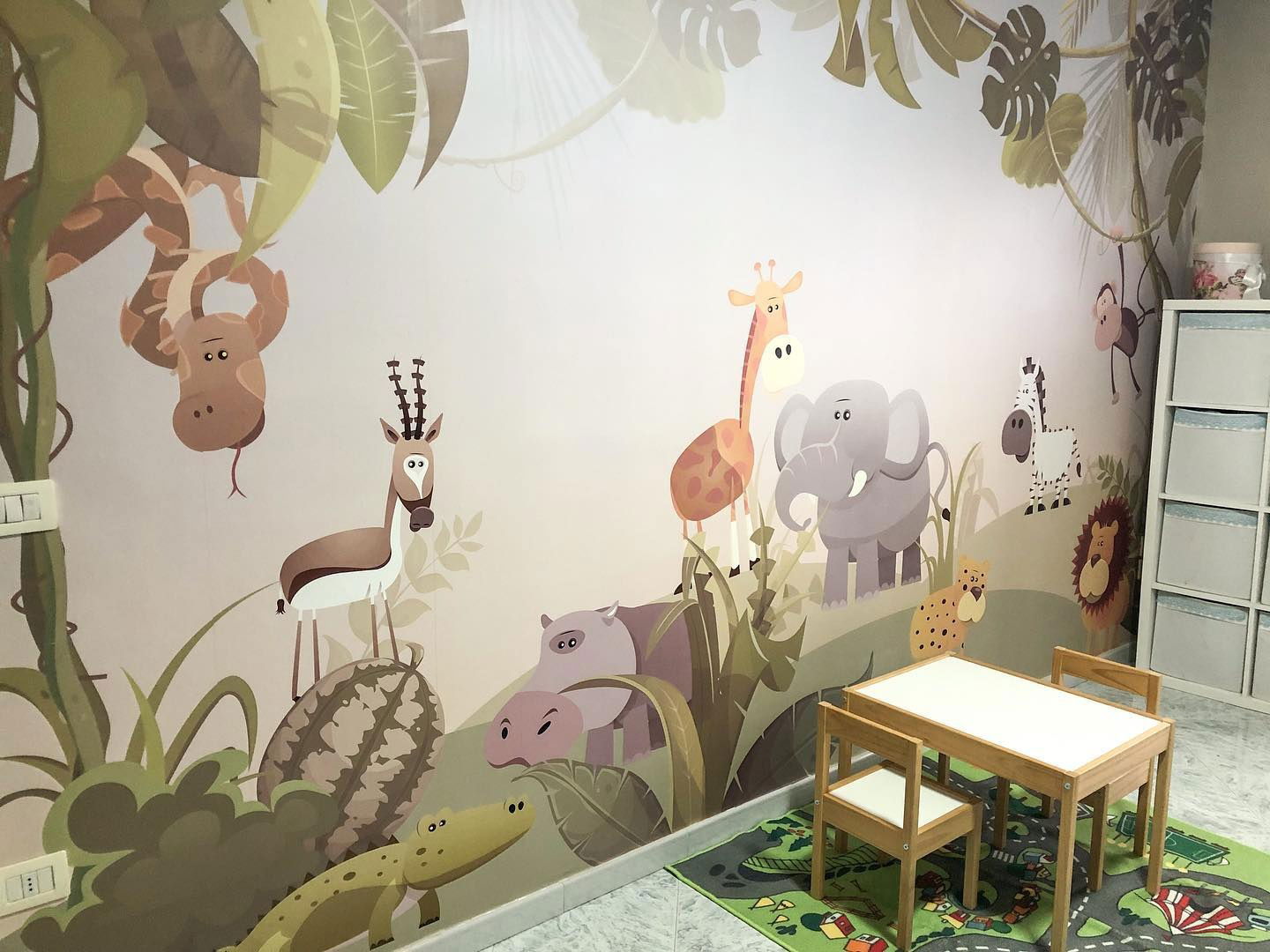 Child's room with jungle animal wall mural, small table and chairs, and green road-themed rug.