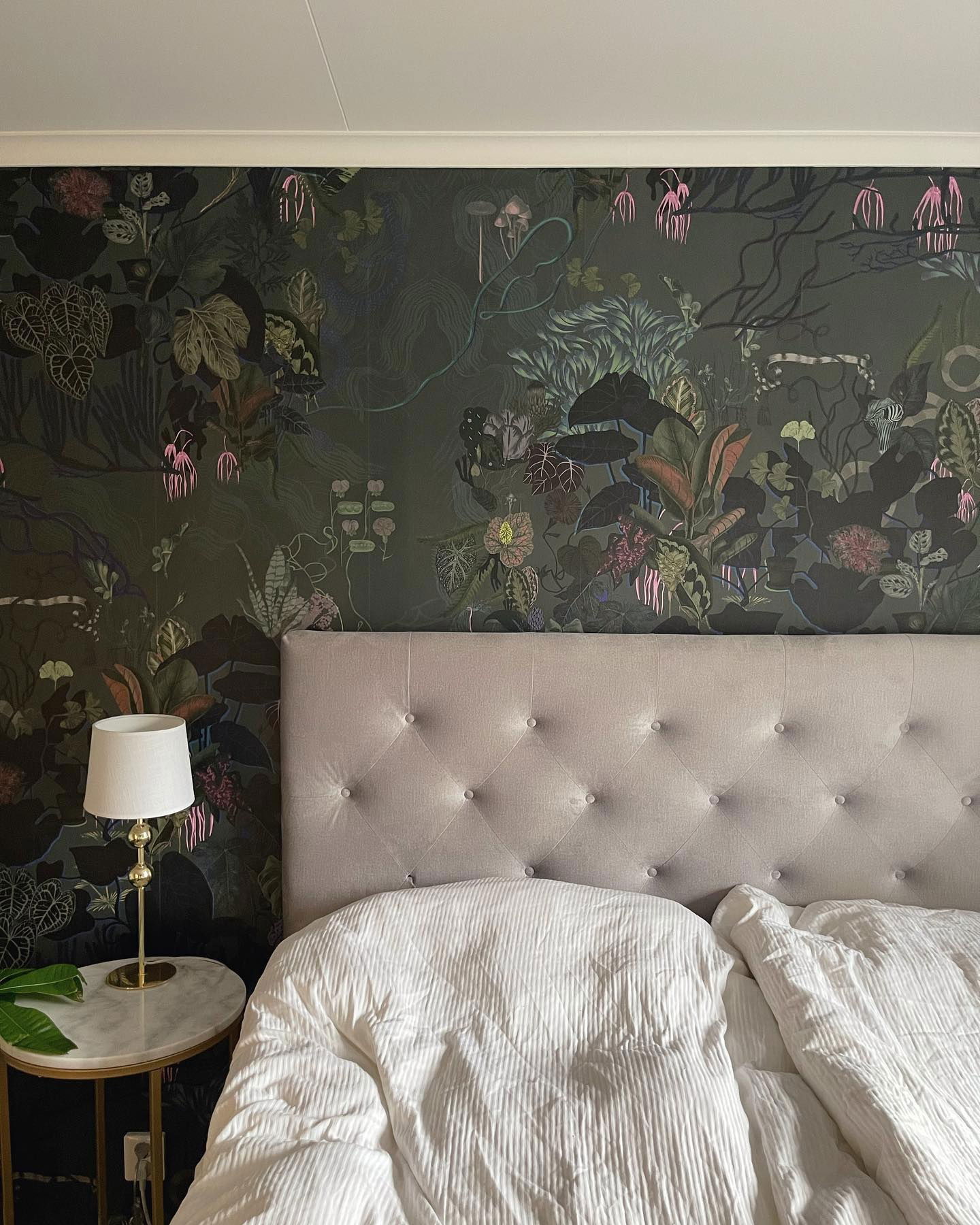 Bedroom with tufted headboard, white bedding, and botanical wallpaper. A lamp sits on a marble-topped side table.