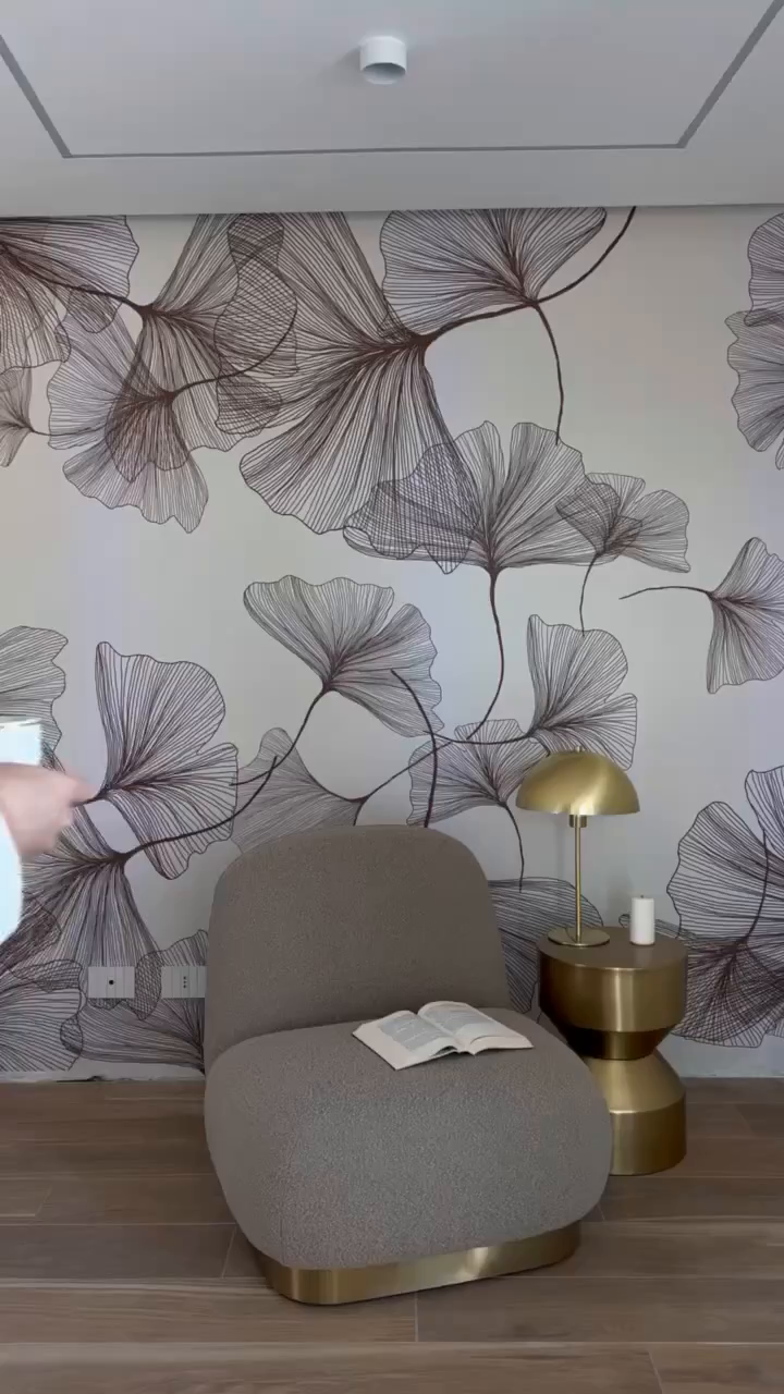 Interior with floral wallpaper, gray chair with book, and gold side table with lamp.