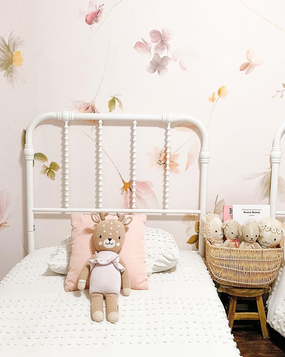 Habitación infantil con cama de metal blanco, papel tapiz floral y peluche de ciervo. Cesta de mimbre con animales de peluche.