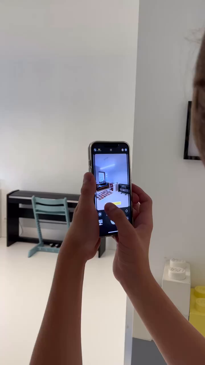 Hands holding a smartphone, taking a photo of a room with a piano and blue chair.