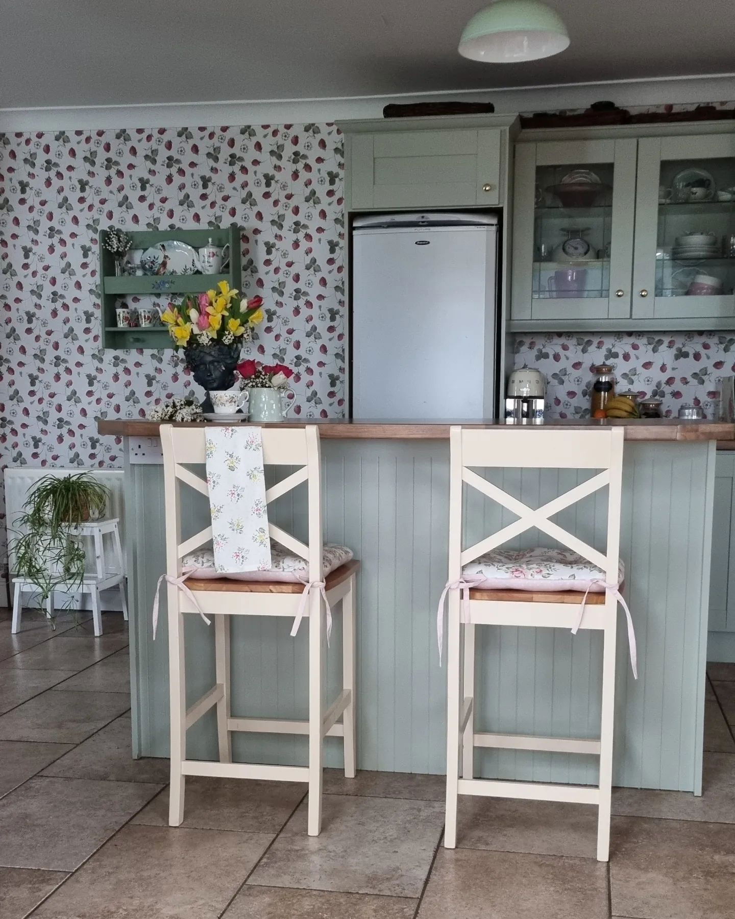 Intérieur de cuisine avec deux tabourets de bar couleur crème, papier peint floral et armoires vert sauge.