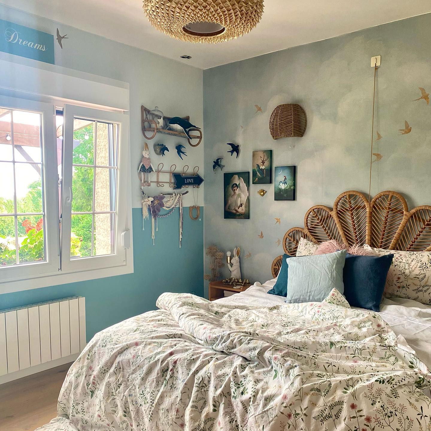 Schlafzimmer mit floraler Bettwäsche, Kopfteil aus Rattan und Wolkenmalerei an den Wänden. Fensterblick ins Grüne.