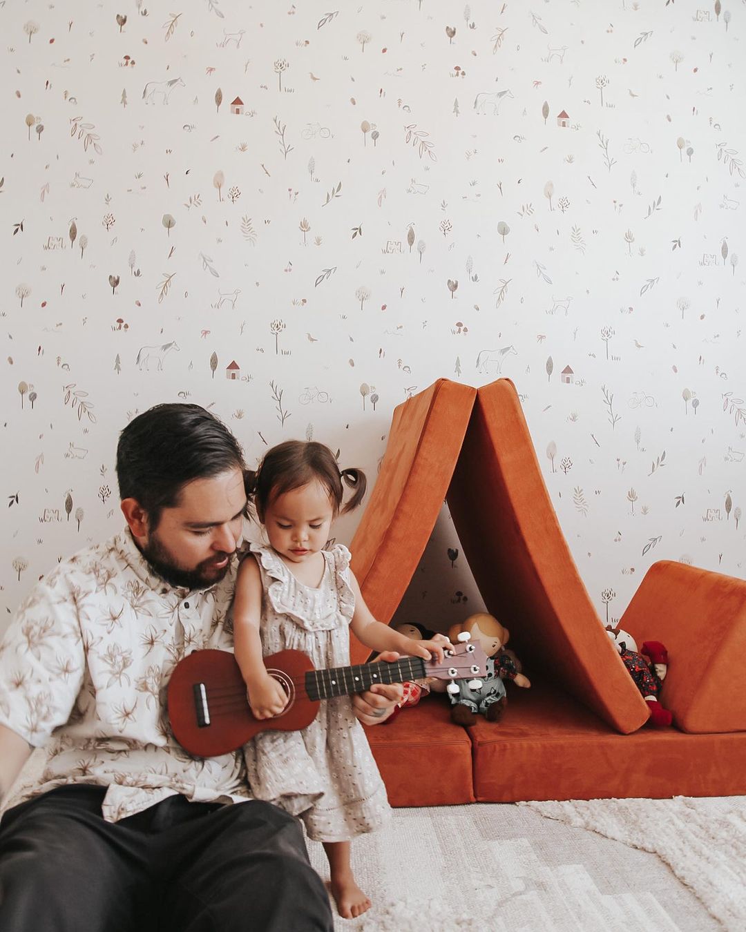 A father and daughter play a ukulele in front of an orange foam play couch.