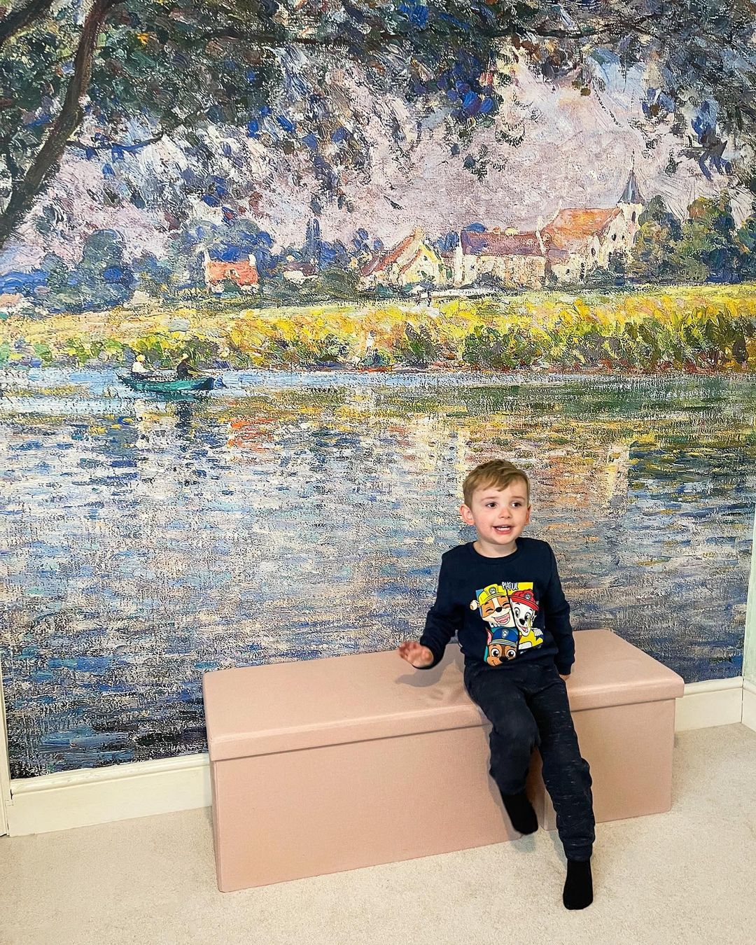 A young boy in a Paw Patrol shirt sits on a pink bench in front of a Monet-style landscape mural.