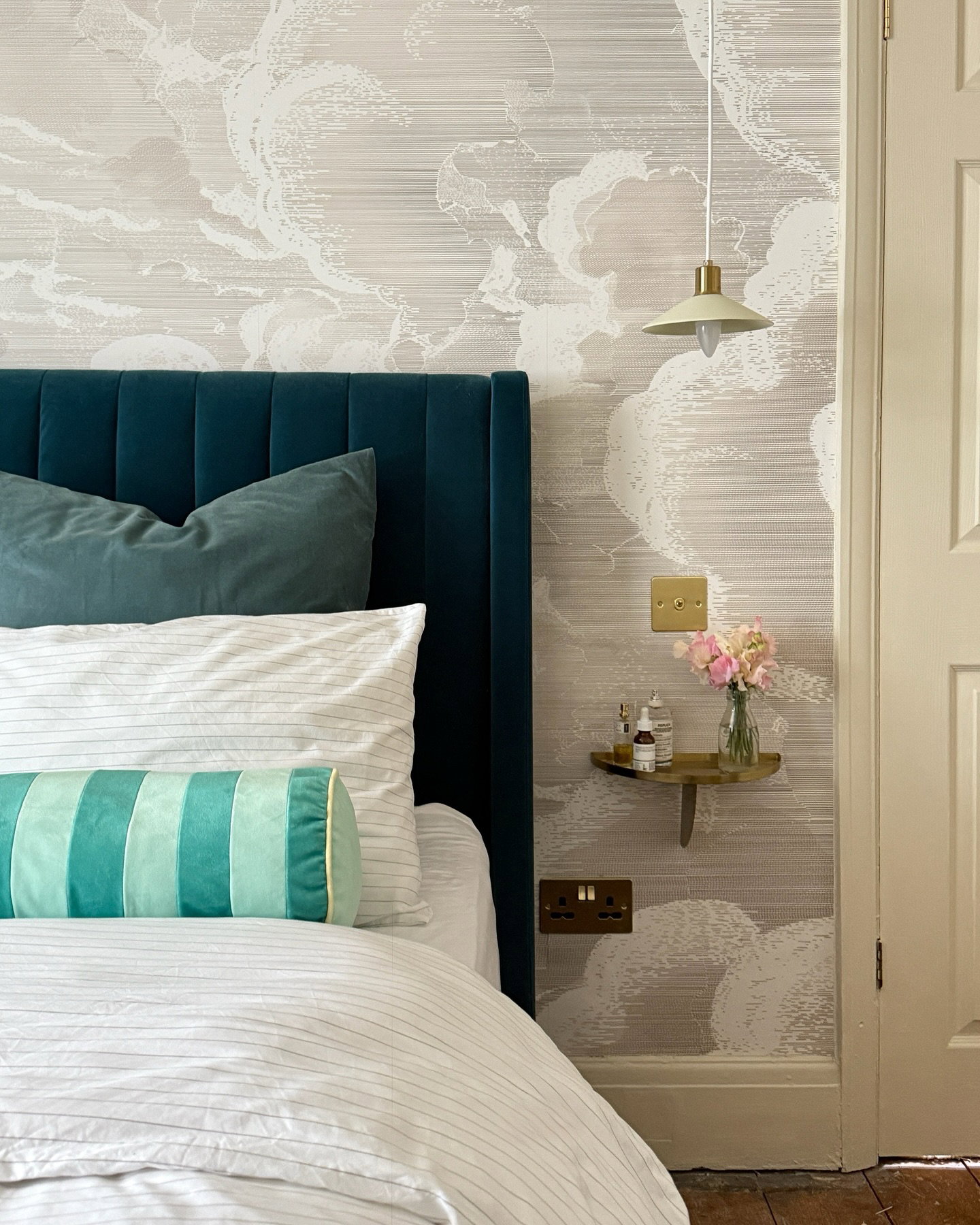 Bedroom with a teal velvet headboard, white striped bedding, and light grey cloud wallpaper. A shelf with pink flowers and a pale green pendant light.