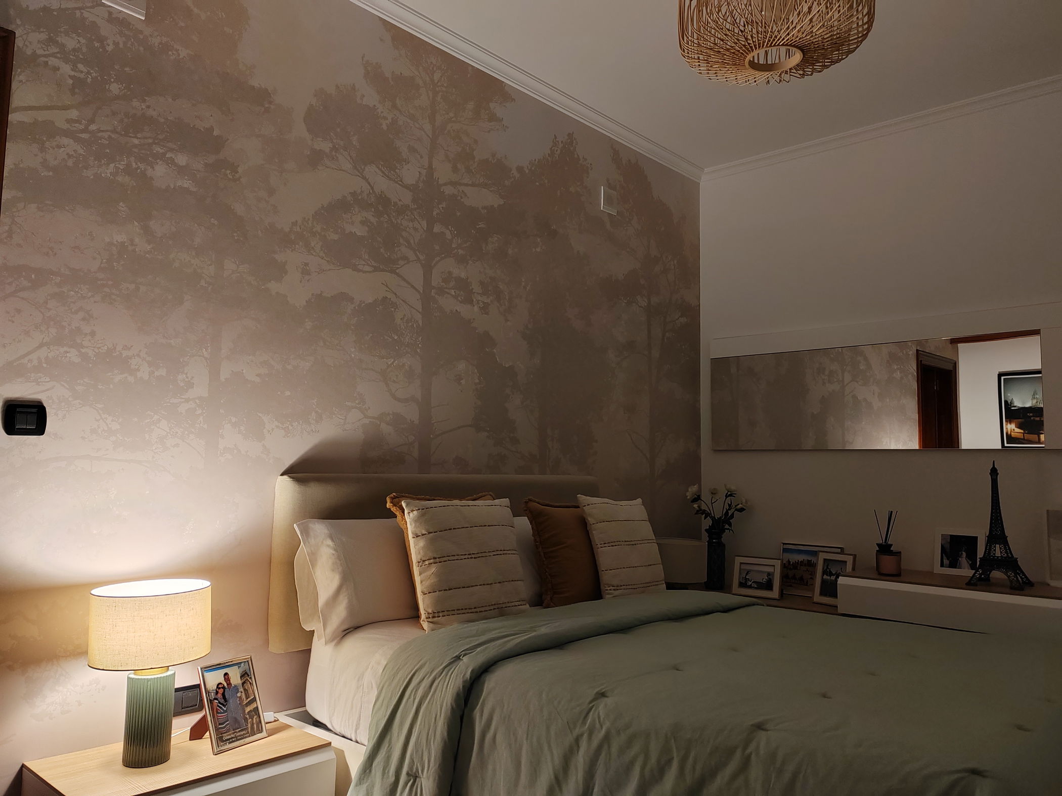 Camera da letto con carta da parati a tema albero, un letto con coperta verde menta e una lampada da comodino.