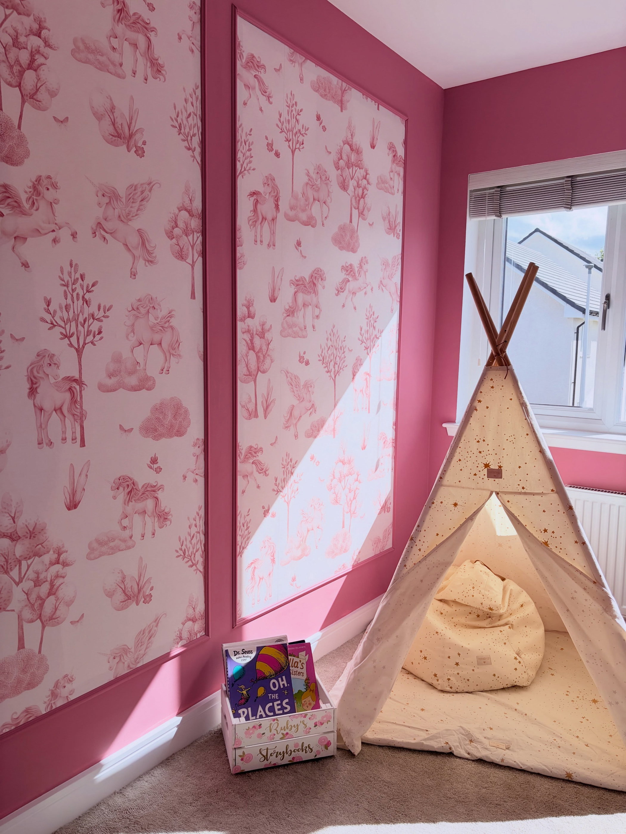 Rosa barnrum med två paneler enhörningstapet, ett krämfärgat tipi-tält med en saccosäck och en låda med sagoböcker.