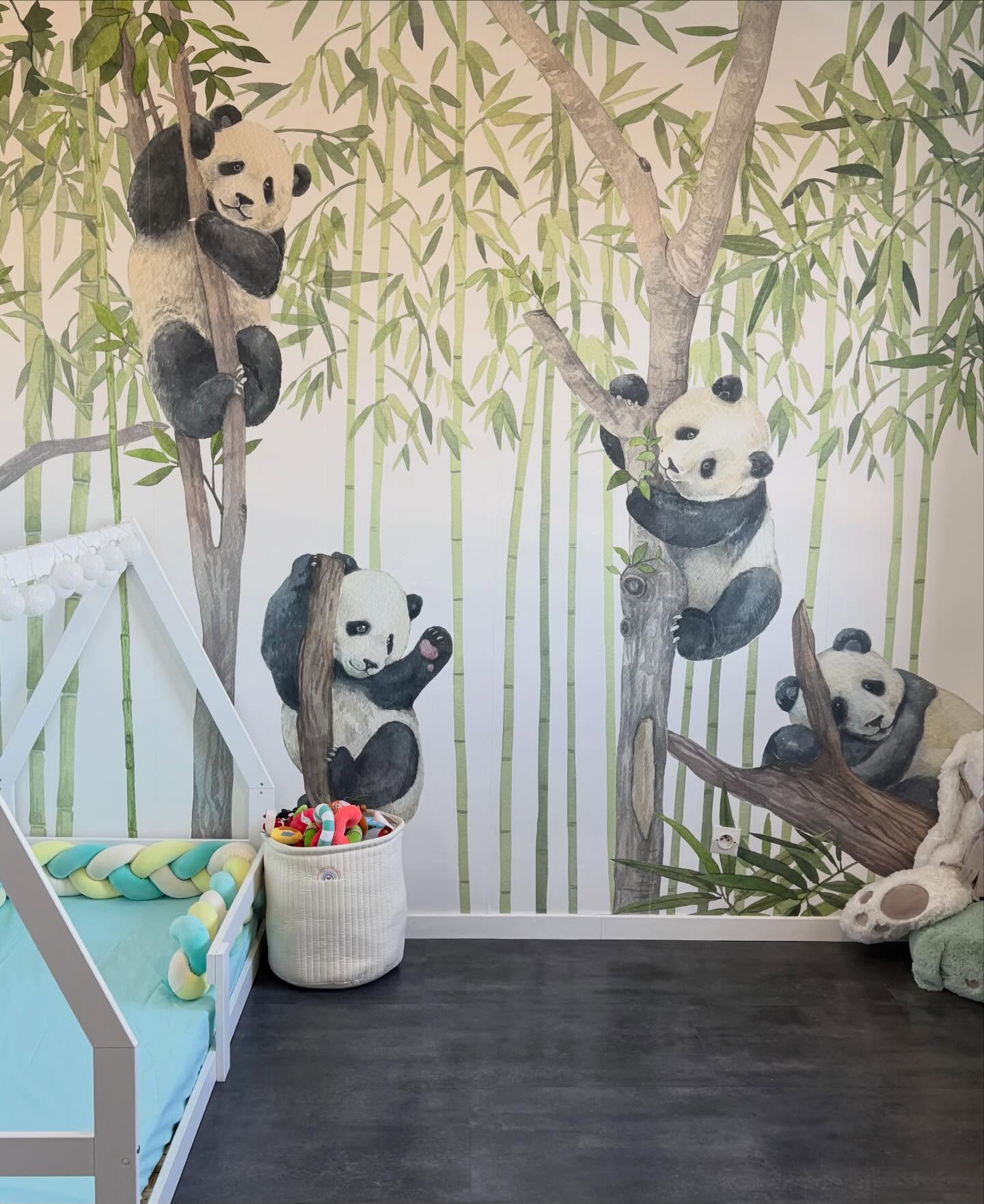 Four pandas on a nursery wall mural, climbing and resting in a bamboo forest. A white toddler bed, toy basket, and stuffed animal are in the room.