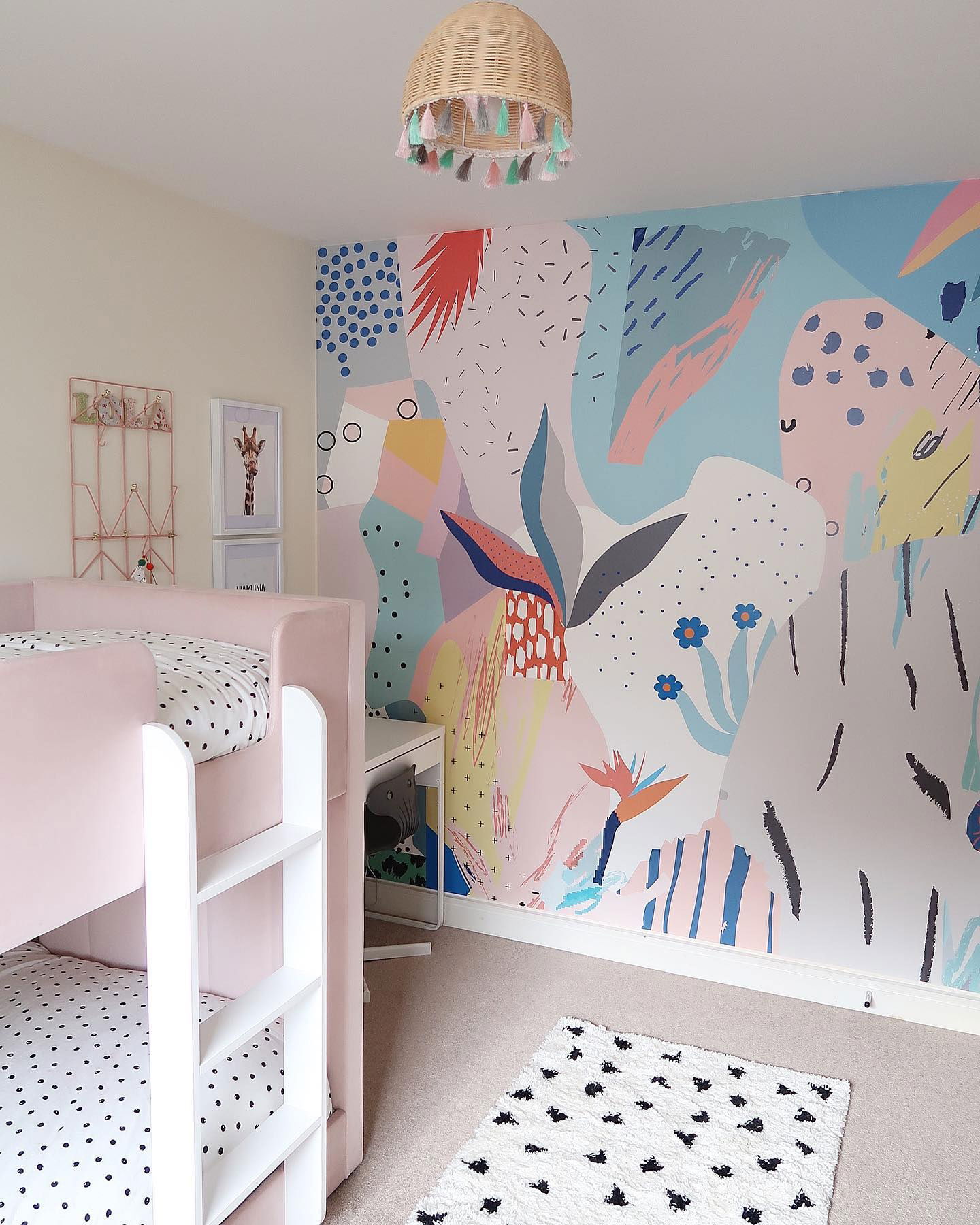 Child's bedroom with pink bunk beds, colorful abstract wallpaper, and a woven tassel light fixture.