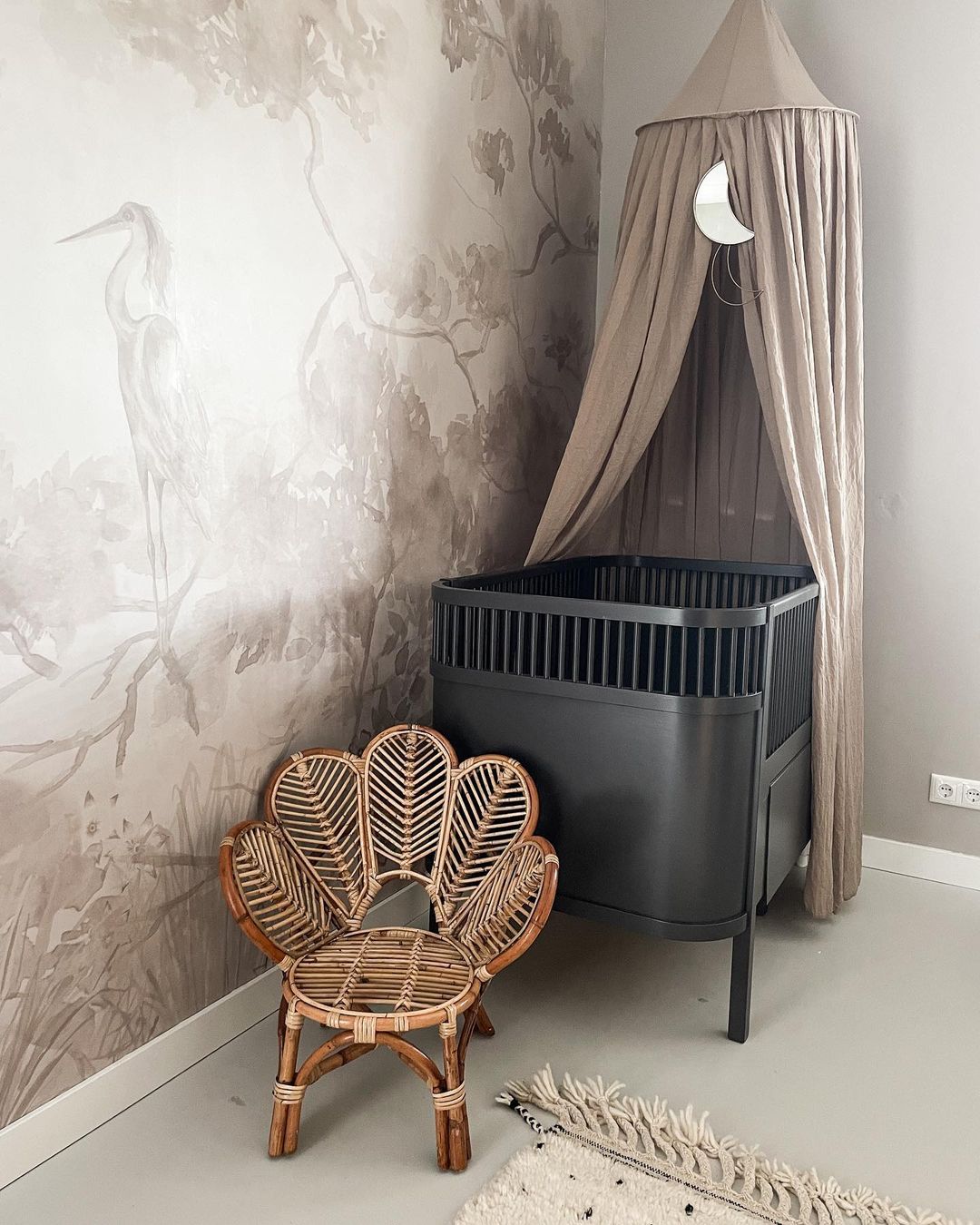Nursery with black crib under beige canopy, flower-shaped rattan chair, and heron wallpaper.