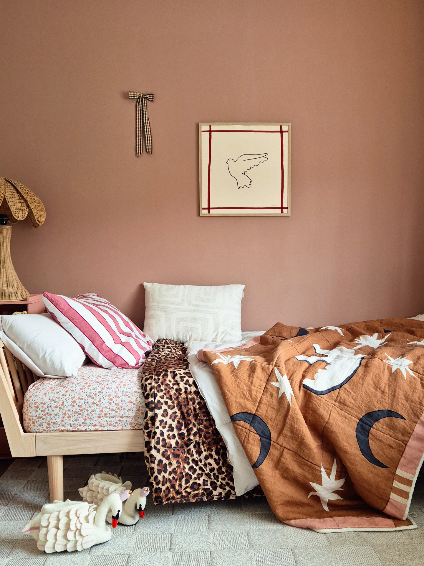 Cama con colcha de luna y estrellas marrón, manta de leopardo y sábanas florales. Pantuflas de cisne sobre alfombra gris. Dibujo de pájaro enmarcado y lazo de vichy en pared rosa empolvado.