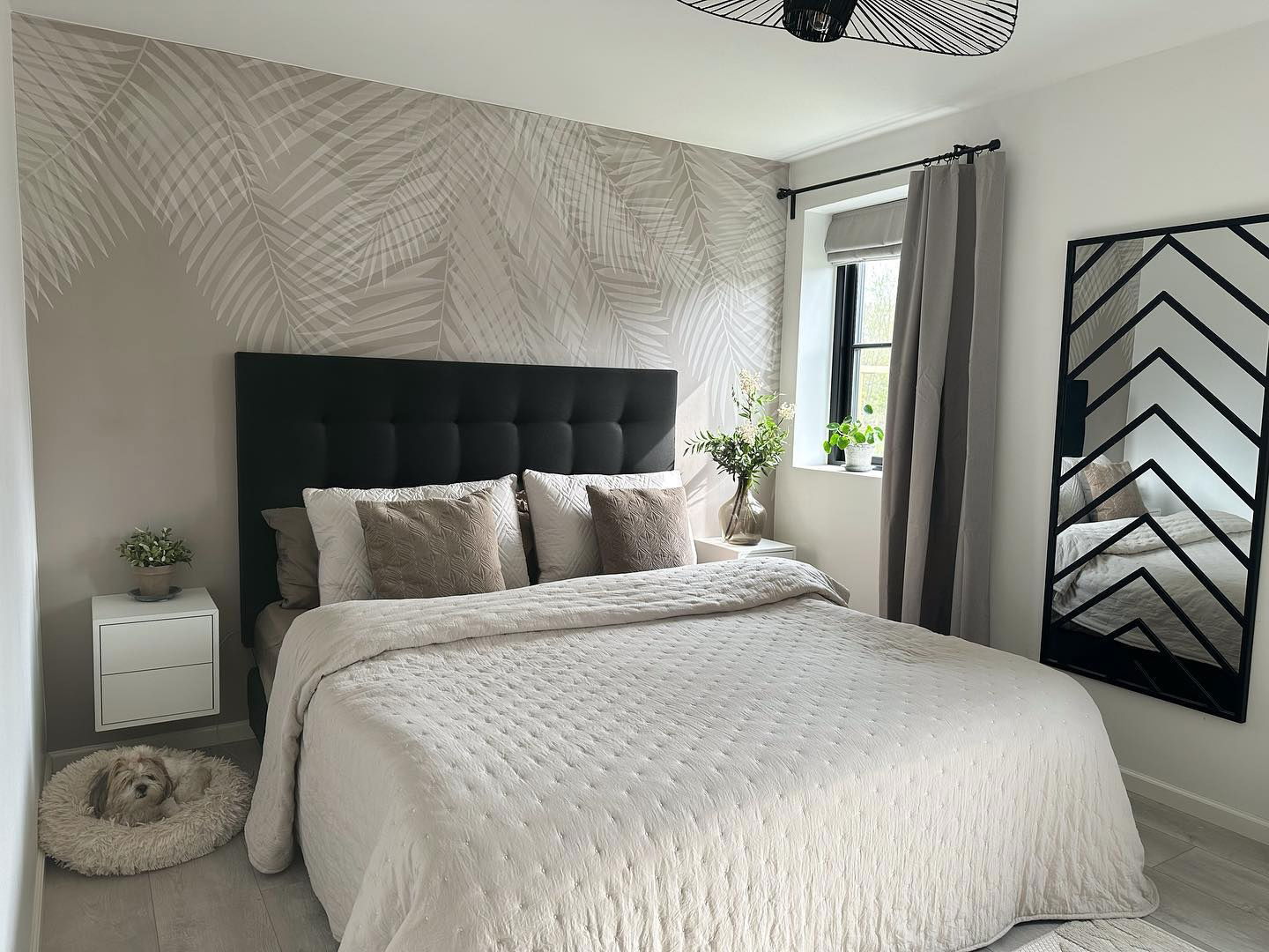 Dormitorio con cabecero negro, colcha beige, papel tapiz de palmeras y cama para perros.