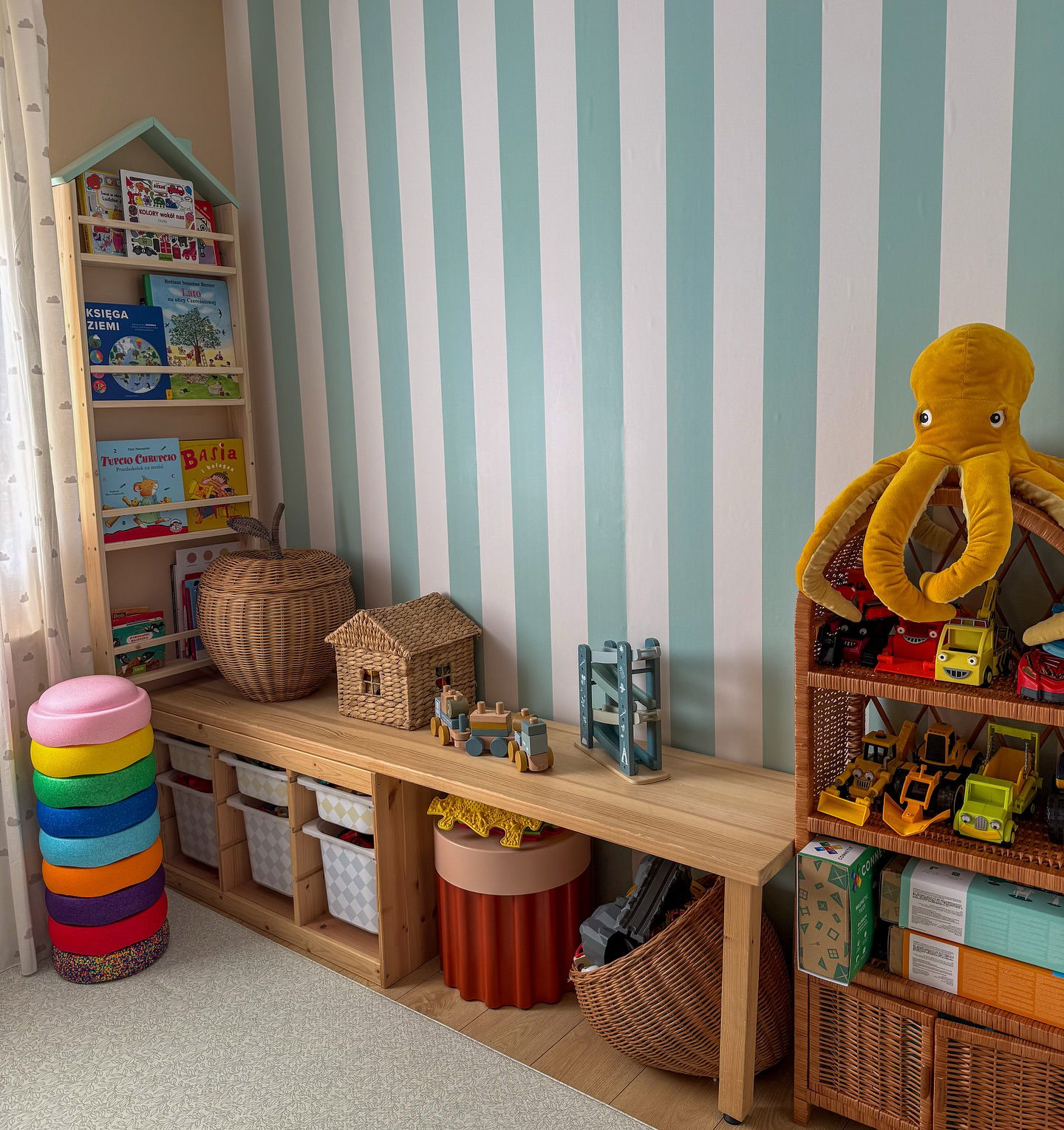 Child's room with wooden shelves, a bench, and a stack of colorful cushions. Plush yellow octopus on shelves with toy vehicles. Striped wallpaper.