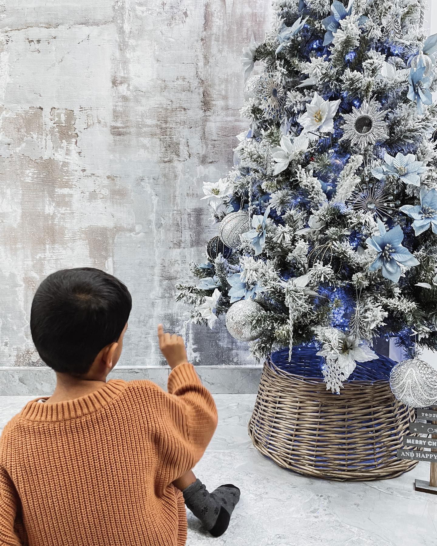 Kind wijst naar een versierde kerstboom met witte en blauwe ornamenten in een geweven mand.