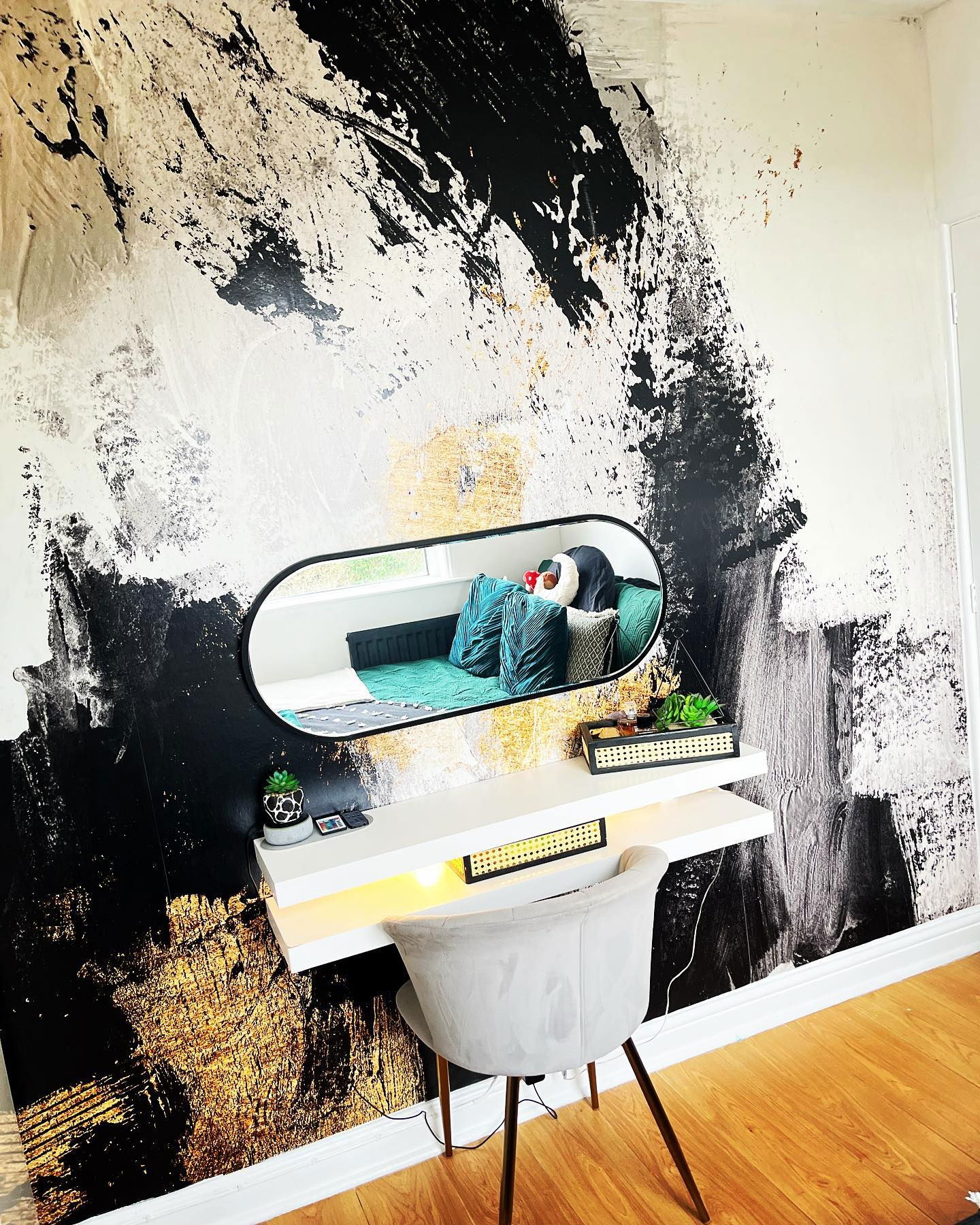 Interior with black, white, and gold abstract wallpaper, floating shelves, oval mirror, and gray chair.