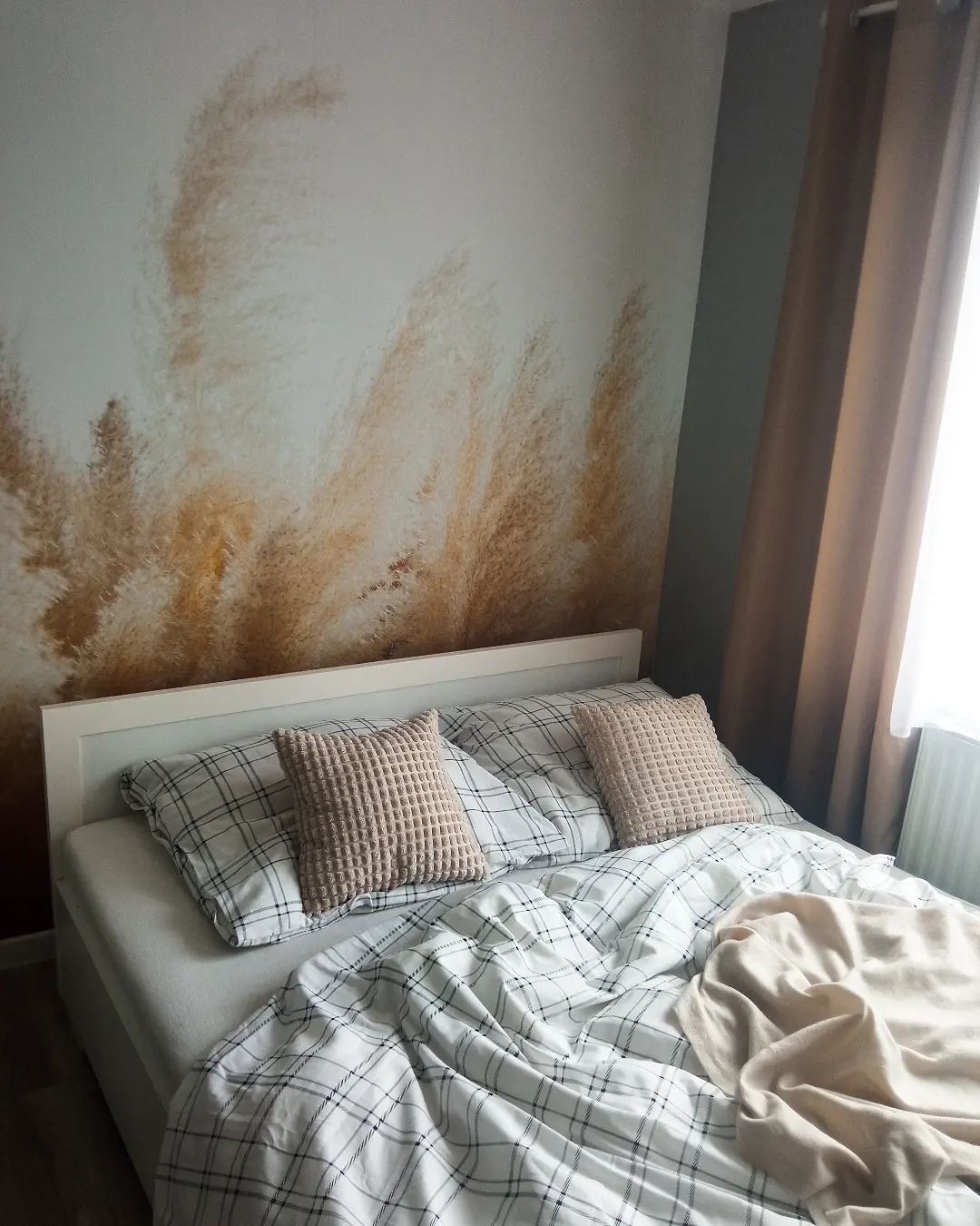 Dormitorio con cama blanca, ropa de cama a cuadros y almohadas color canela. La pared tiene un diseño de hierba de pampa.