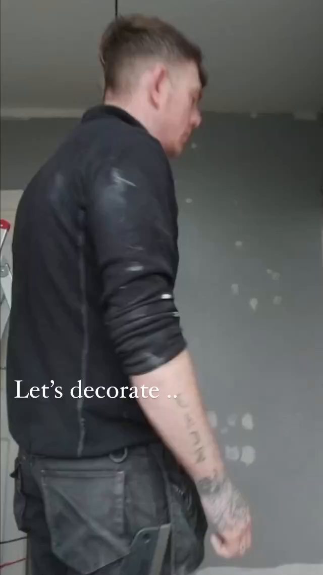 A tattooed man in black work clothes stands in front of a gray wall with white patches. Text overlay: "Let's decorate..."