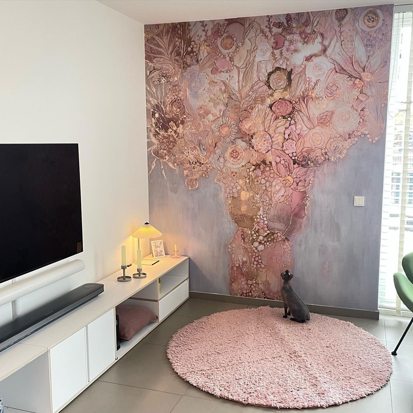 Stue med blomstermalerier, TV og rosa lurvete teppe. En grå katt sitter på teppet.
