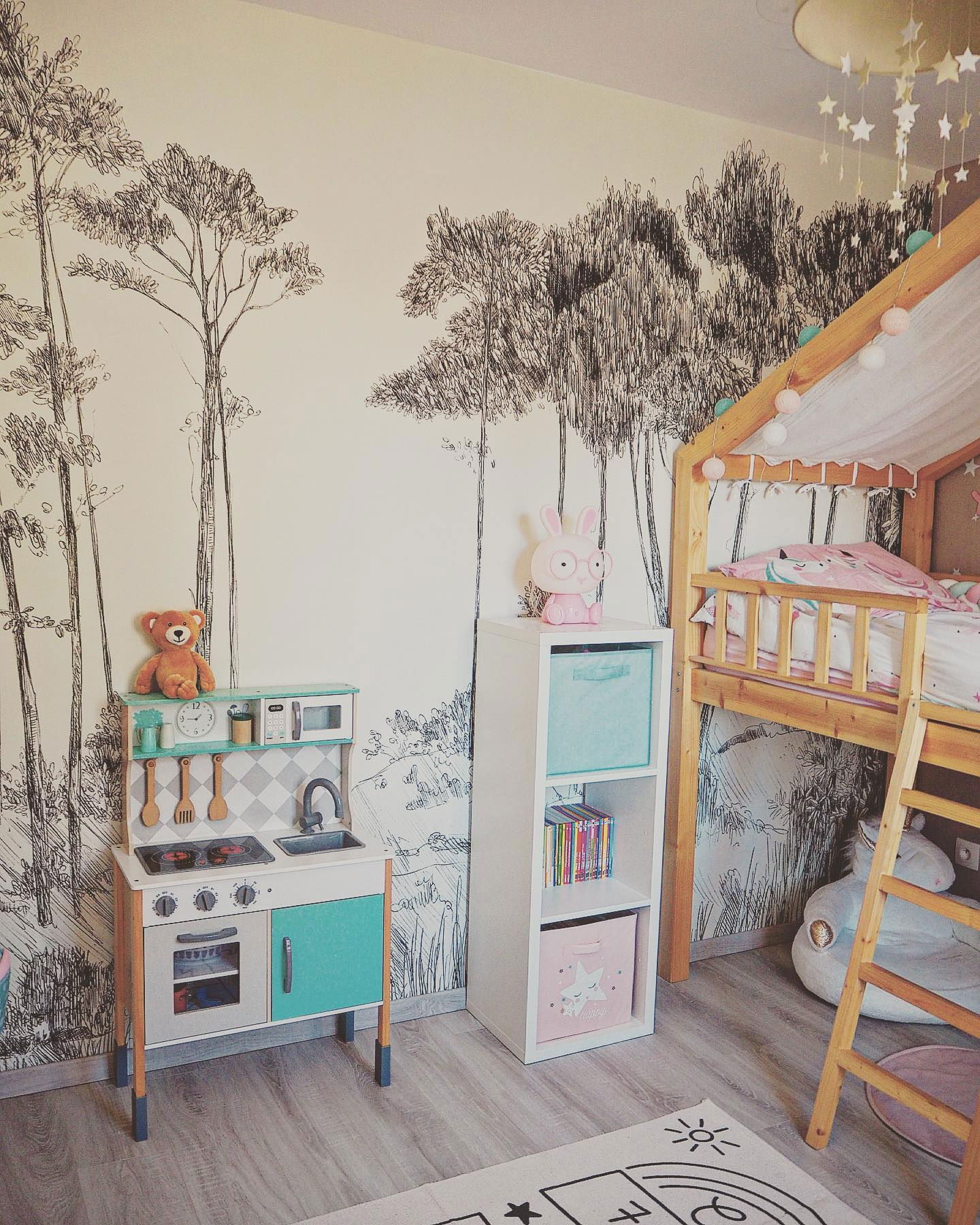 Kinderzimmer mit Baumtapete, hölzerner Spielküche, weißem Bücherregal und Hochbett mit Lichterkette.