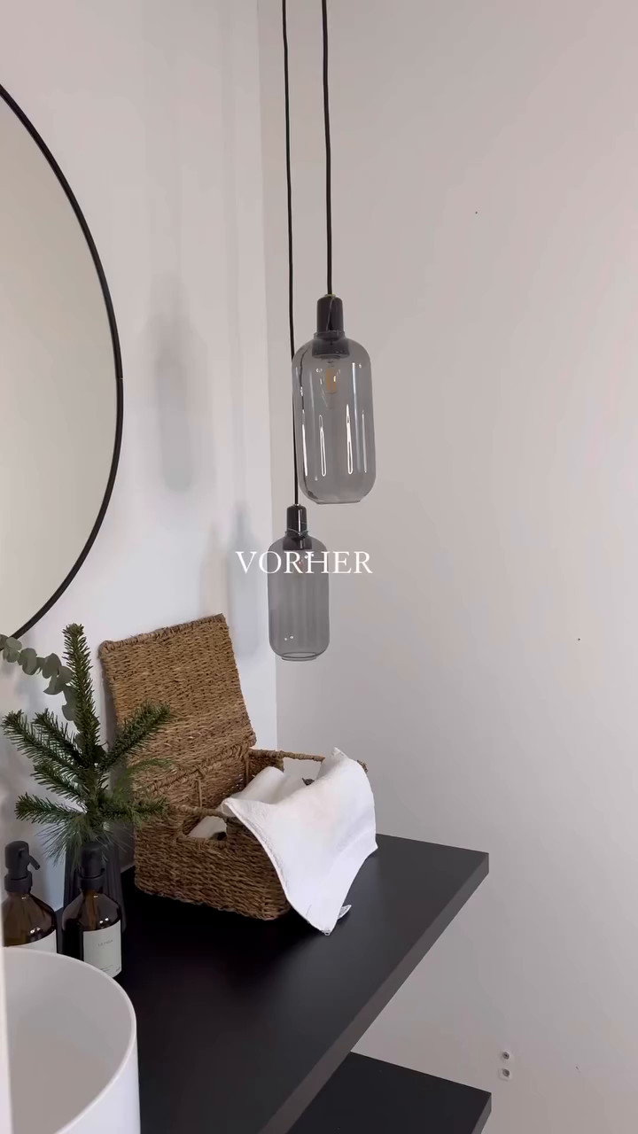 Bathroom corner with gray pendant lights, round mirror, and woven basket on a black shelf.