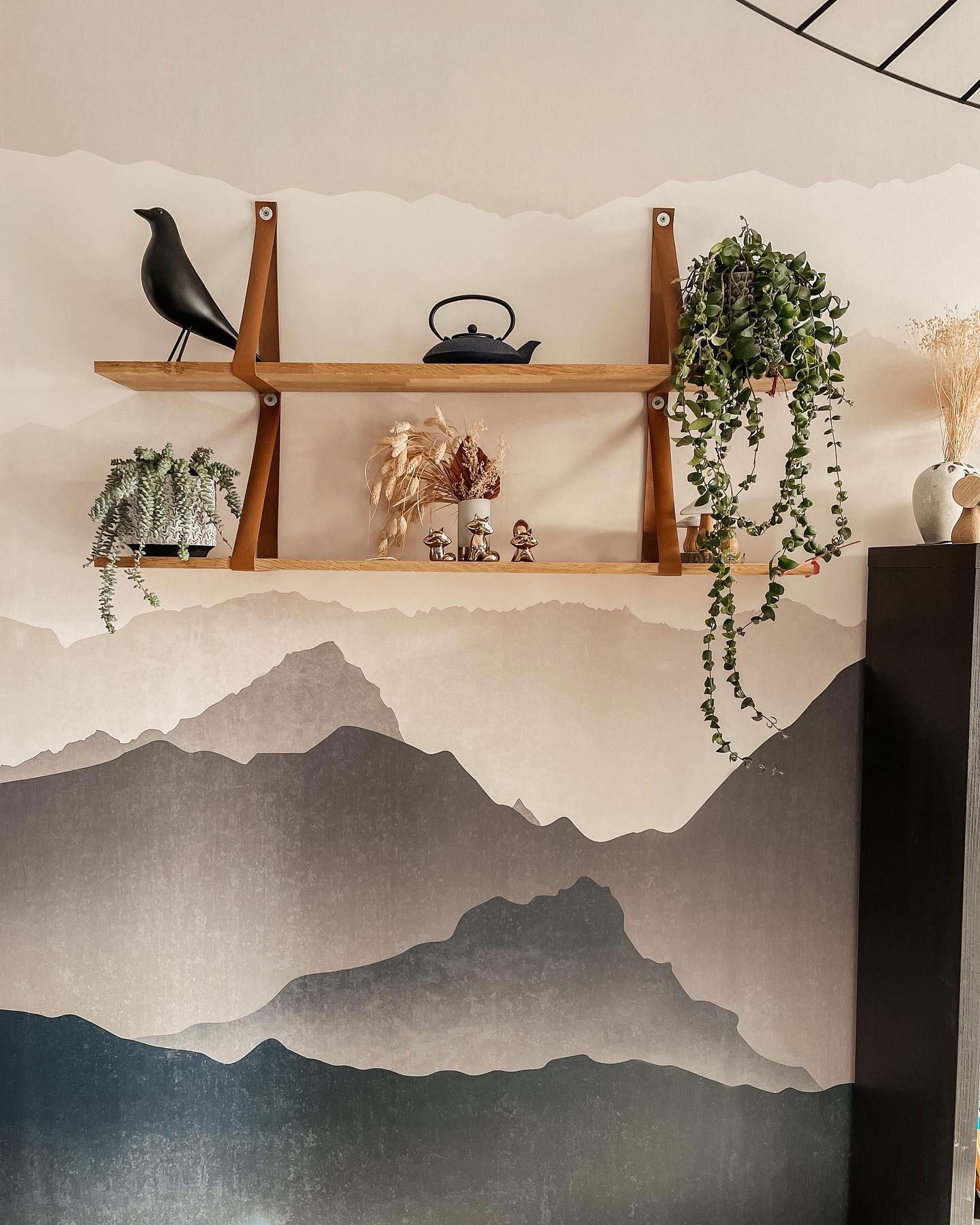 Two-tiered wooden shelf with leather straps, against a mountain mural. Shelf holds plants, teapot, and decorative objects.