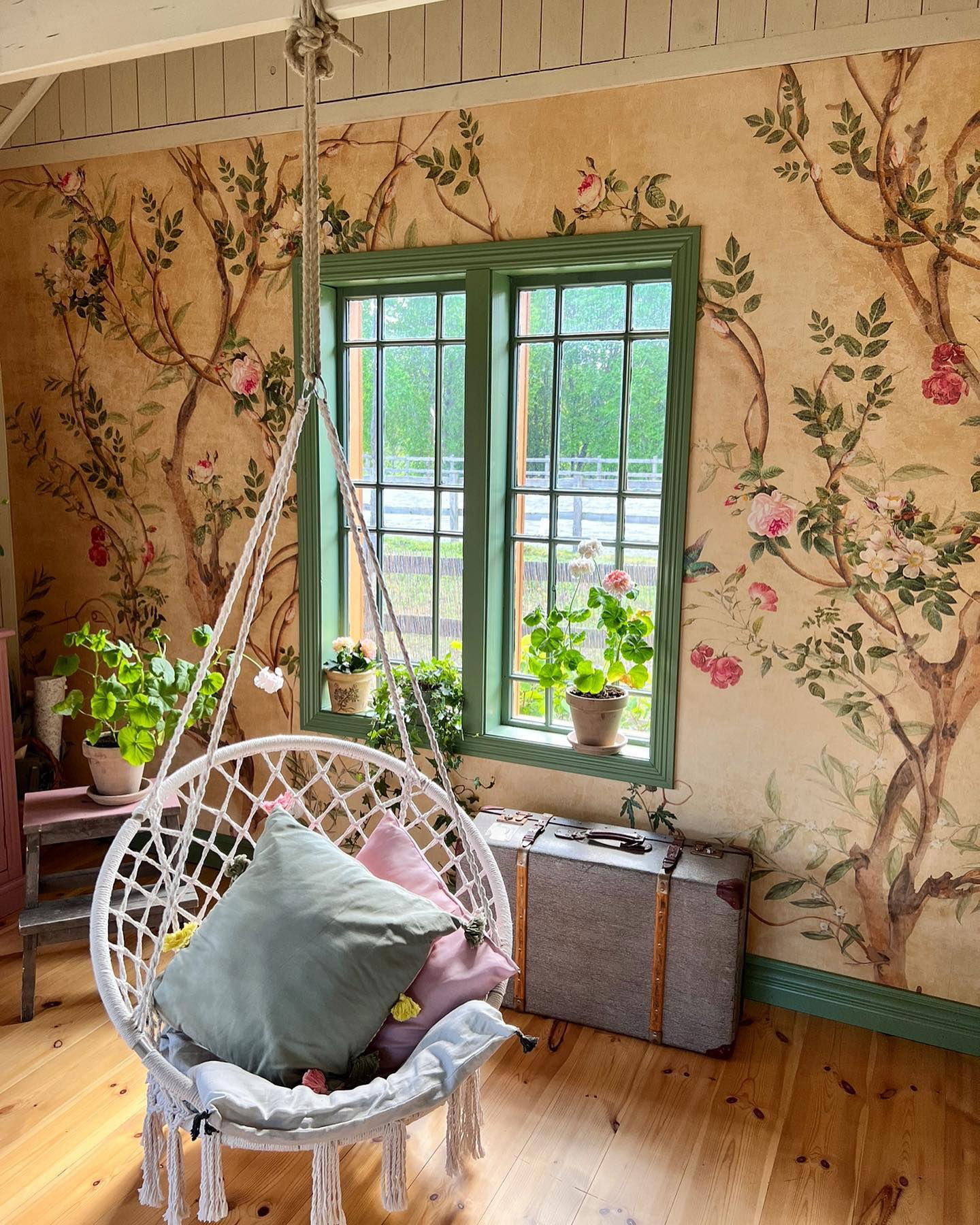 Hyggeligt værelse med blomstret tapet, grønt vindue og hængende macrame-stol med puder.