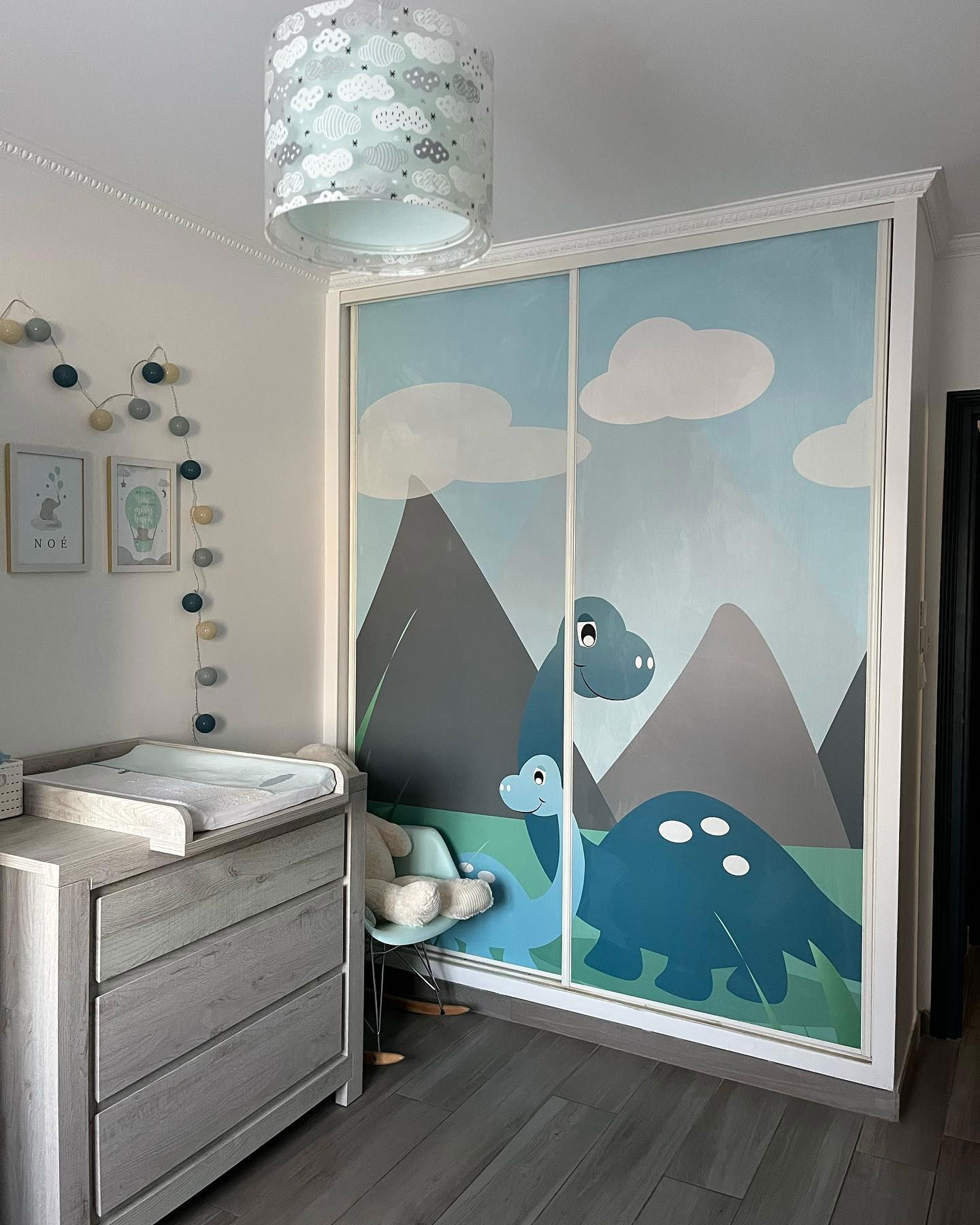 Kinderzimmer mit grauer Kommode, Wolkenlampe und Dinosaurier-Wandbild auf den Schranktüren.