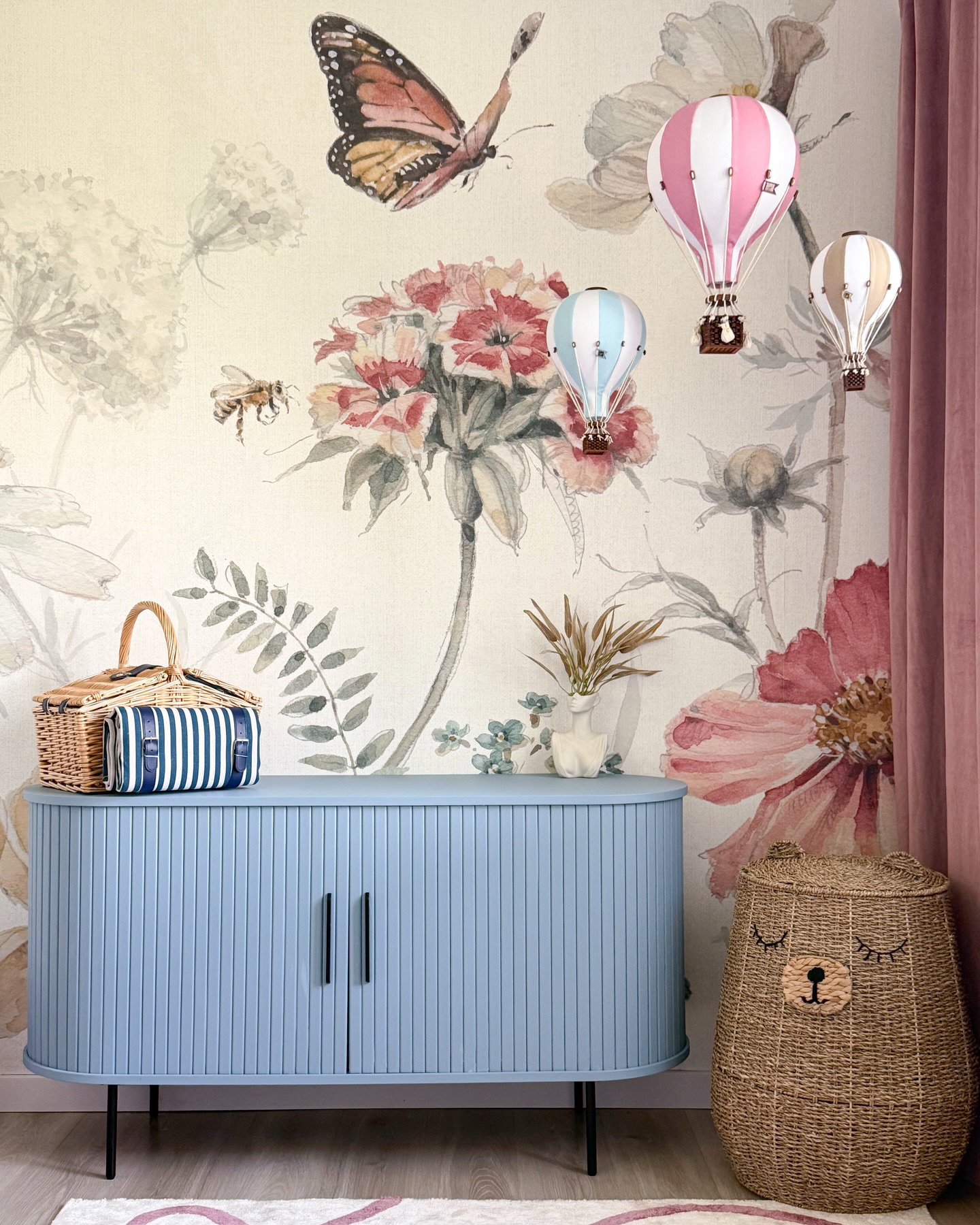 Blaue Kommode mit Picknickkorb und geflochtenem Bärenkorb vor floraler Tapete mit Heißluftballons.