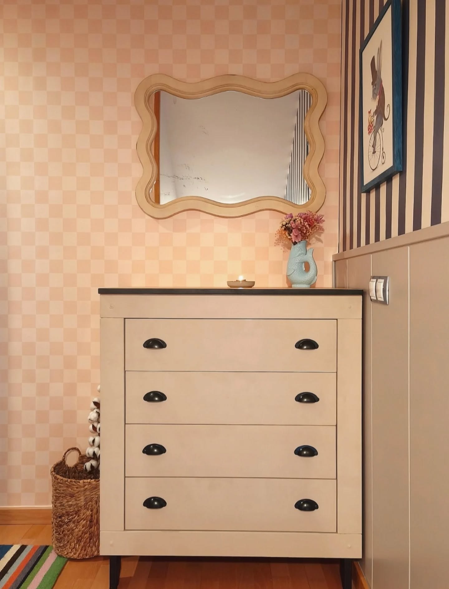 Light beige dresser with black hardware below a scalloped mirror and a framed picture on a striped wall.
