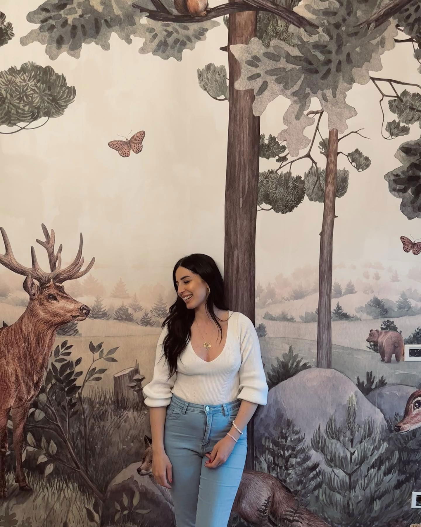 A woman in a white sweater and jeans poses in front of a forest mural. The mural features deer, bears, and trees.