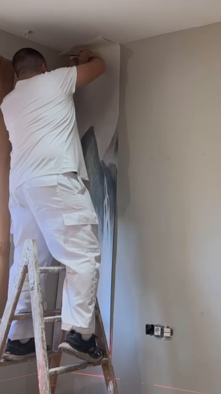Man on ladder installing mountain mural wallpaper.