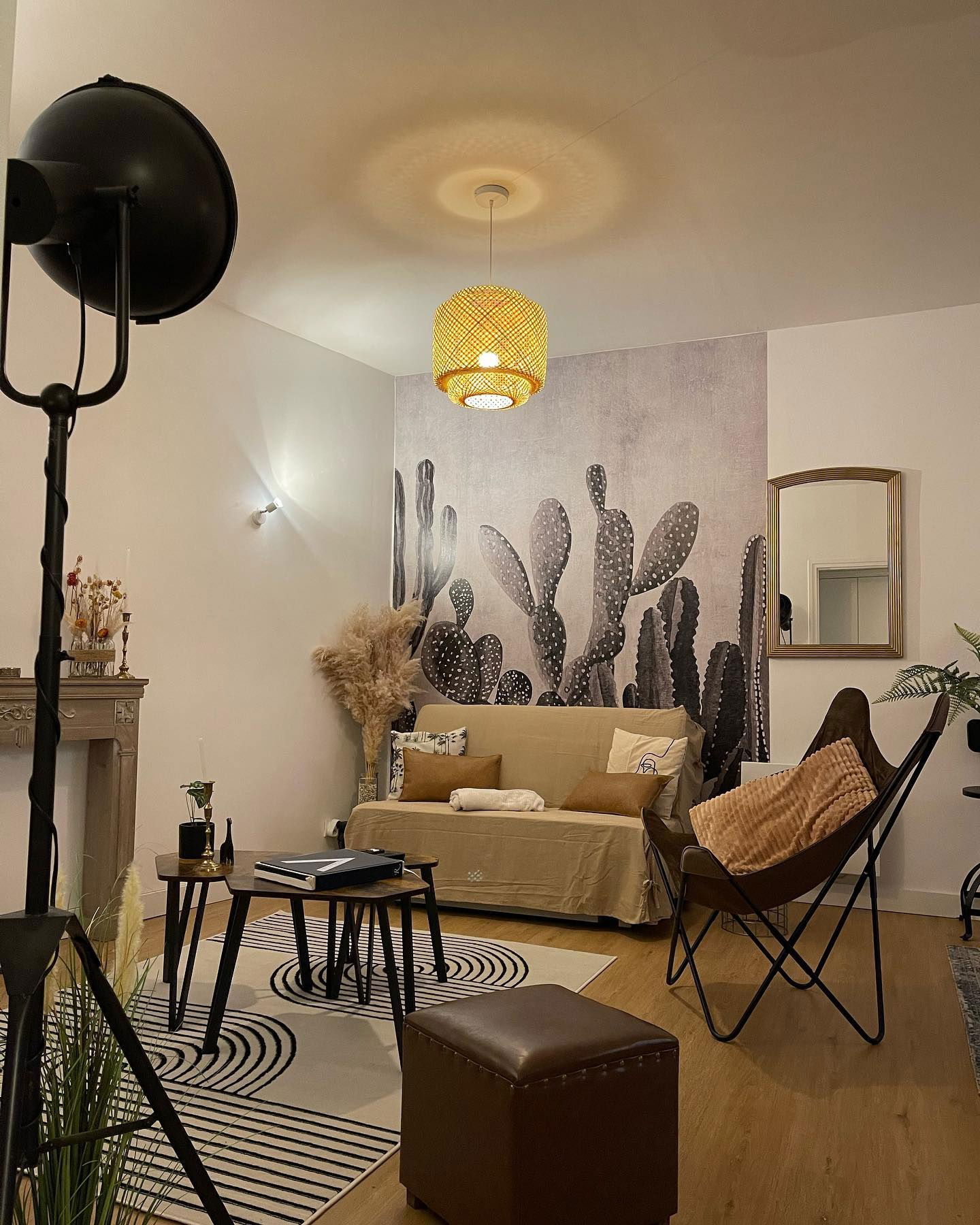 Gemütliches Wohnzimmer mit beigem Sofa, Kaktus-Wandbild und Schmetterlingsstuhl. Gewebte Hängeleuchte und geometrischer Teppich.