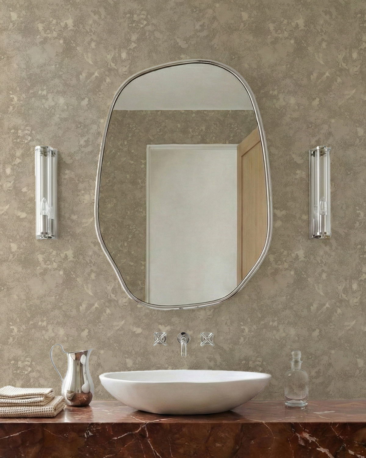 Miroir de forme irrégulière au-dessus d'un lavabo blanc sur un comptoir en marbre avec pichet argenté et serviettes.
