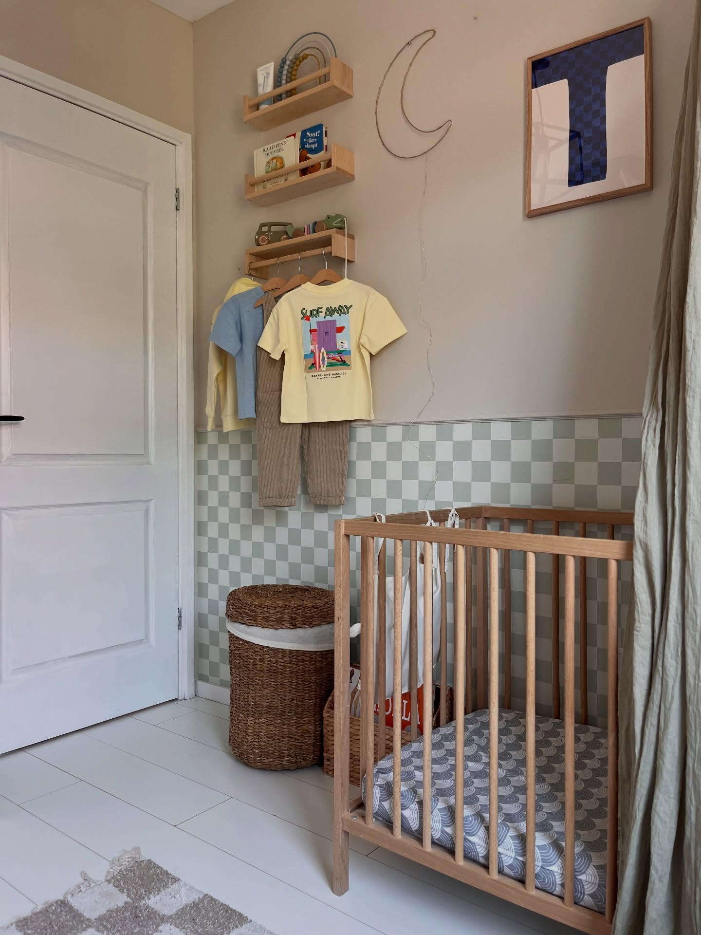 En lys tre-seng på et barnerom med rutete tapet. Trehyller inneholder bøker og leker, med barneklær hengende under. En månelampe og innrammet kunst pryder veggen.