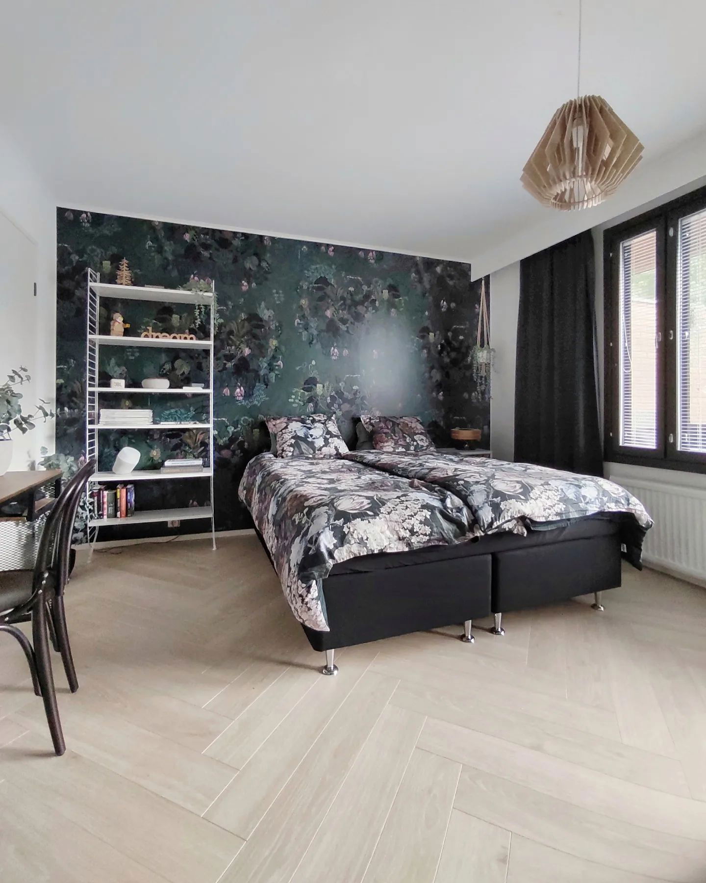Chambre avec papier peint floral, cadre de lit noir et planchers de bois clair.