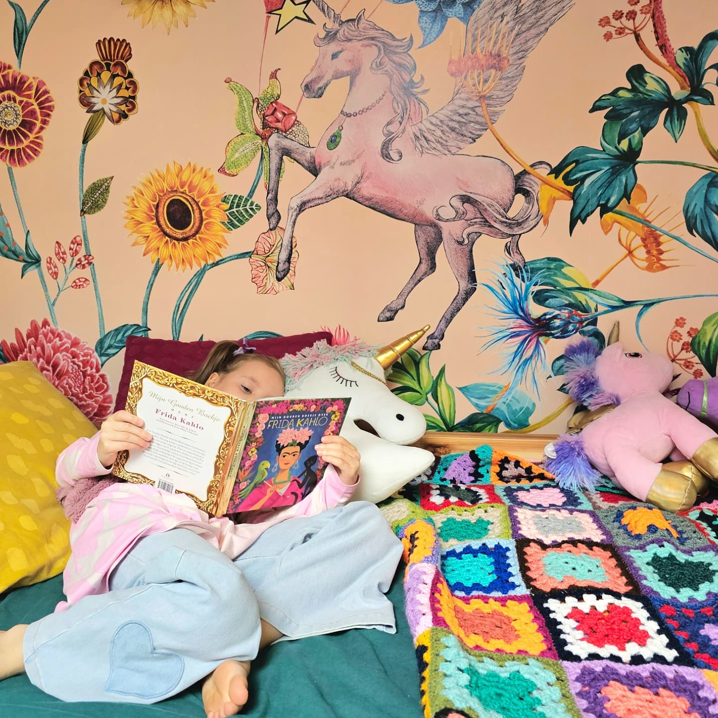 Dziewczyna czyta książkę "Frida Kahlo" w łóżku z tapetą w jednorożce i kocem z szydełka.