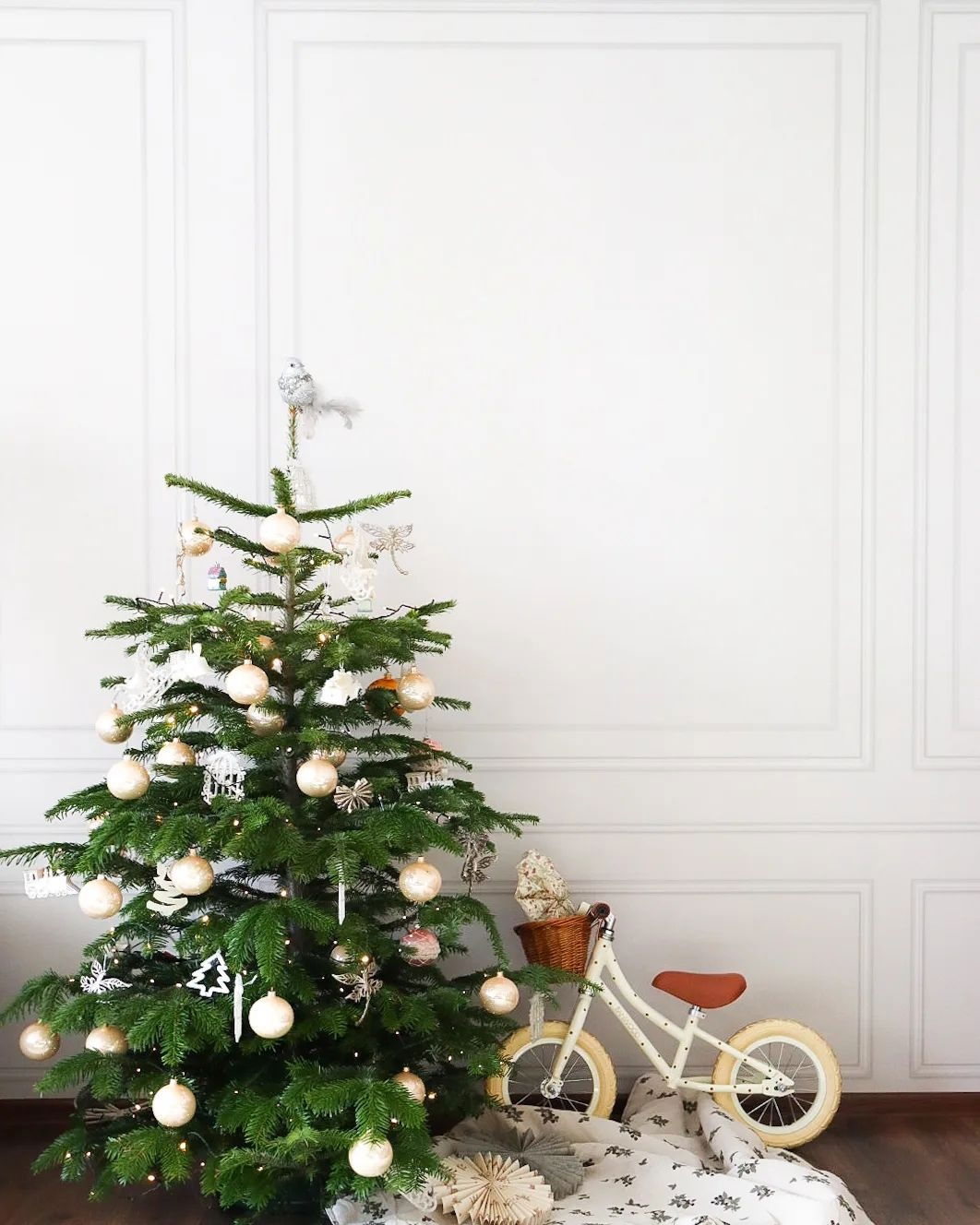 Decorated Christmas tree with white balance bike and basket in front of white paneled wall.