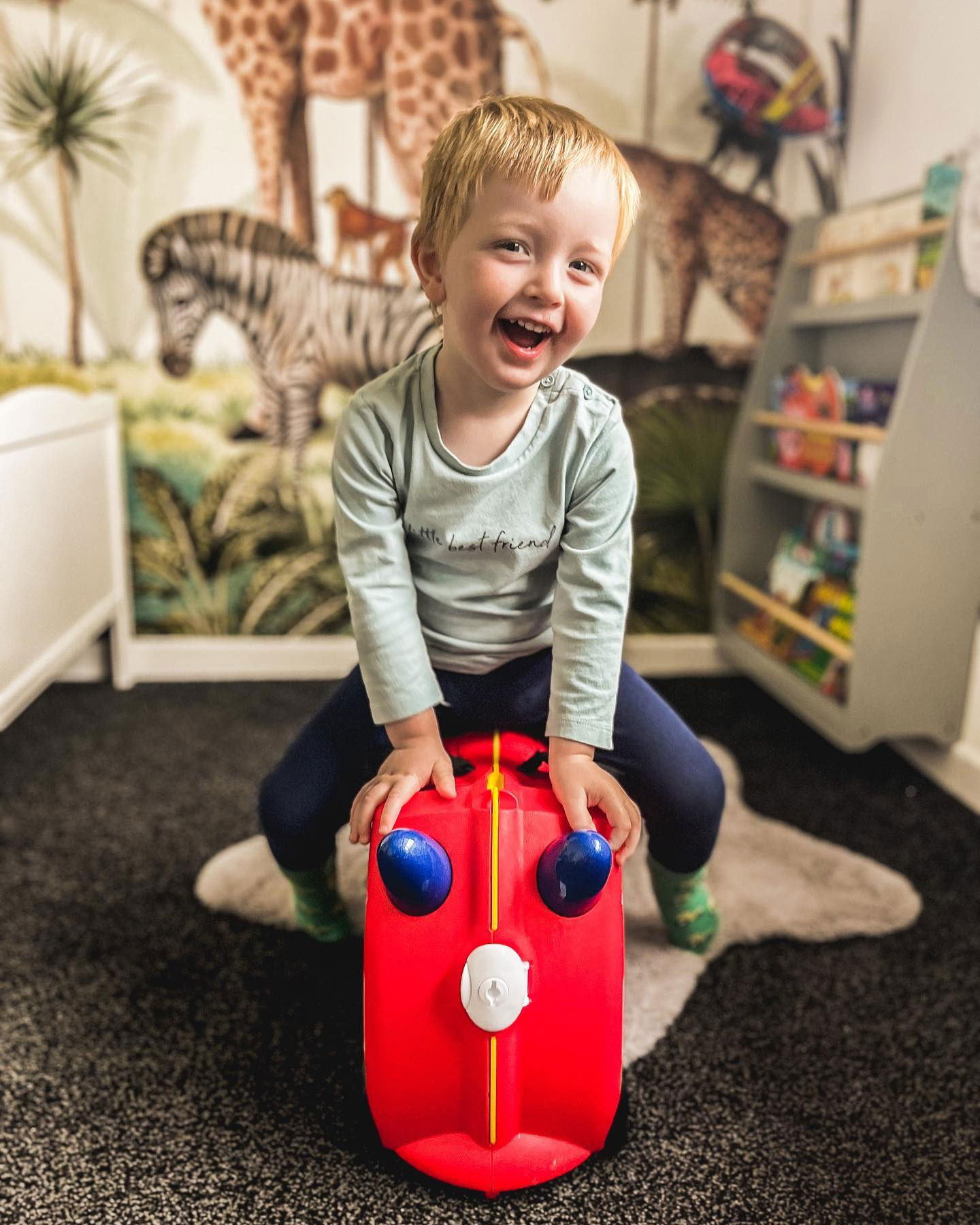 Smilende småbarn som rir på en rød Wheely Bug-leke i et barnerom.