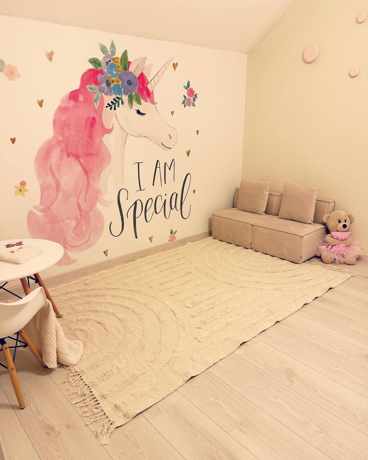 Kinderzimmer mit Einhorn-Wandbild, beigem Teppich, modularem Sofa und Teddybär.