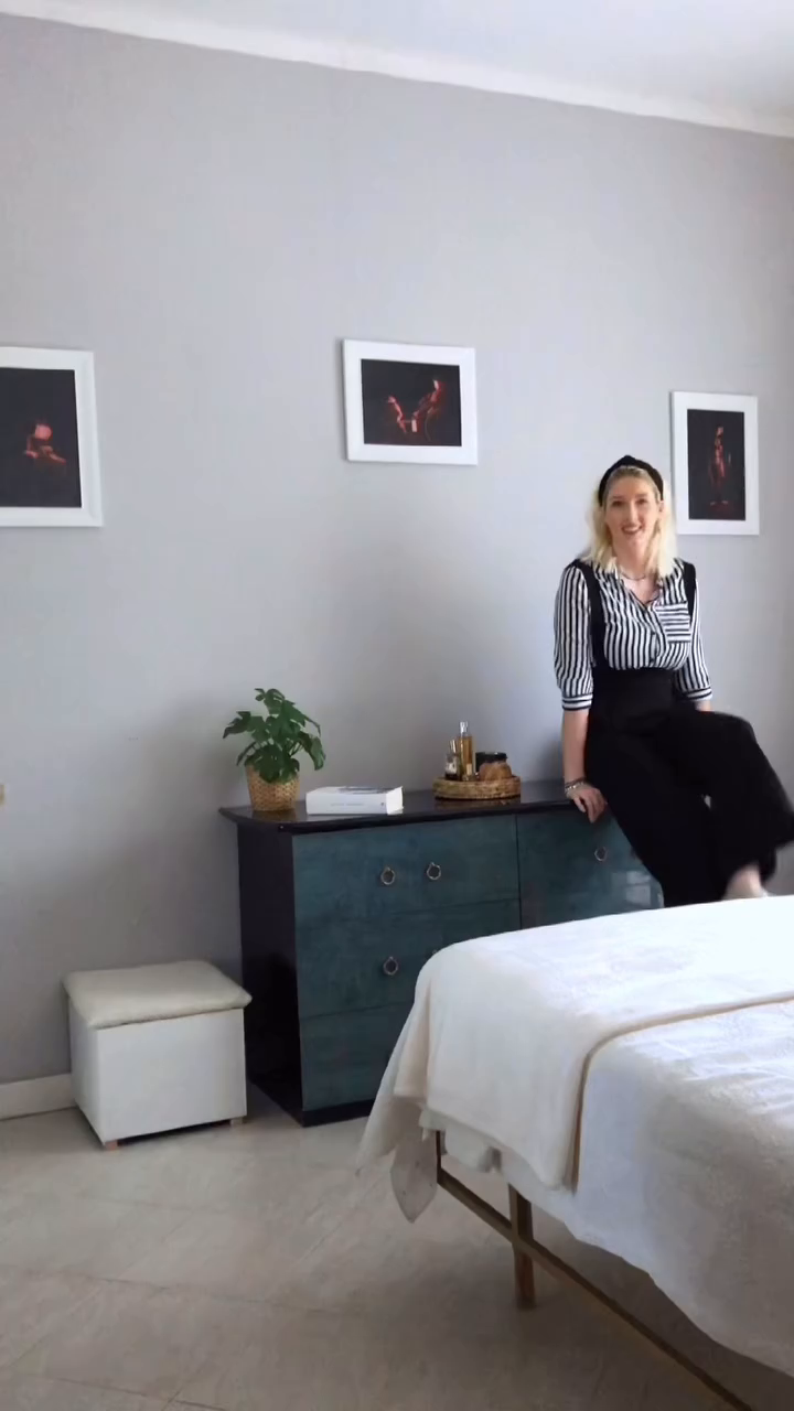 A woman in a striped shirt and black overalls sits on a blue dresser in a spa room.