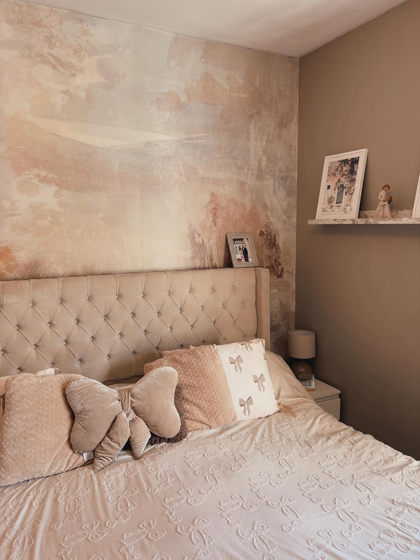 Light beige tufted bed with bow pillows and embroidered duvet. Abstract beige and pink mural on wall behind. Right wall has a shelf with framed photos.