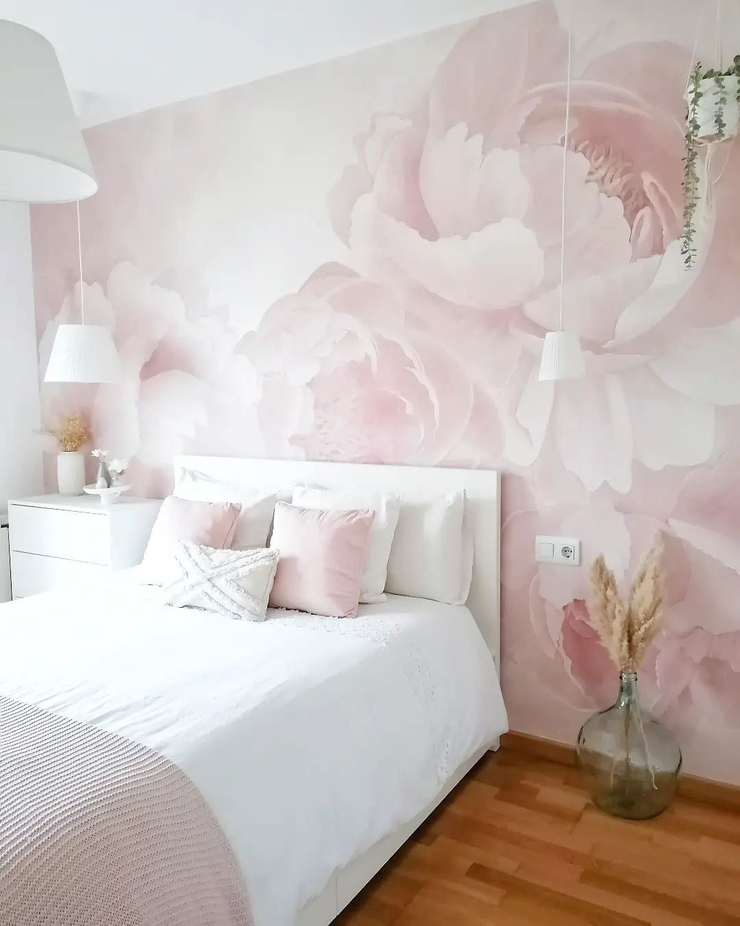 Bedroom with white bedding, pink pillows, and floral wallpaper. Dried pampas grass in a glass vase sits on the wood floor.