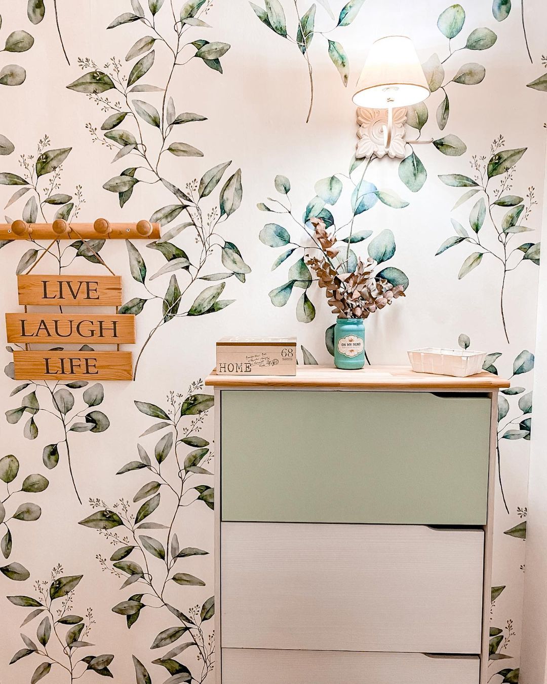 Soveværelsesinteriør med bladagtigt tapet, mintgrøn kommode og "Live Laugh Life"-skilt.