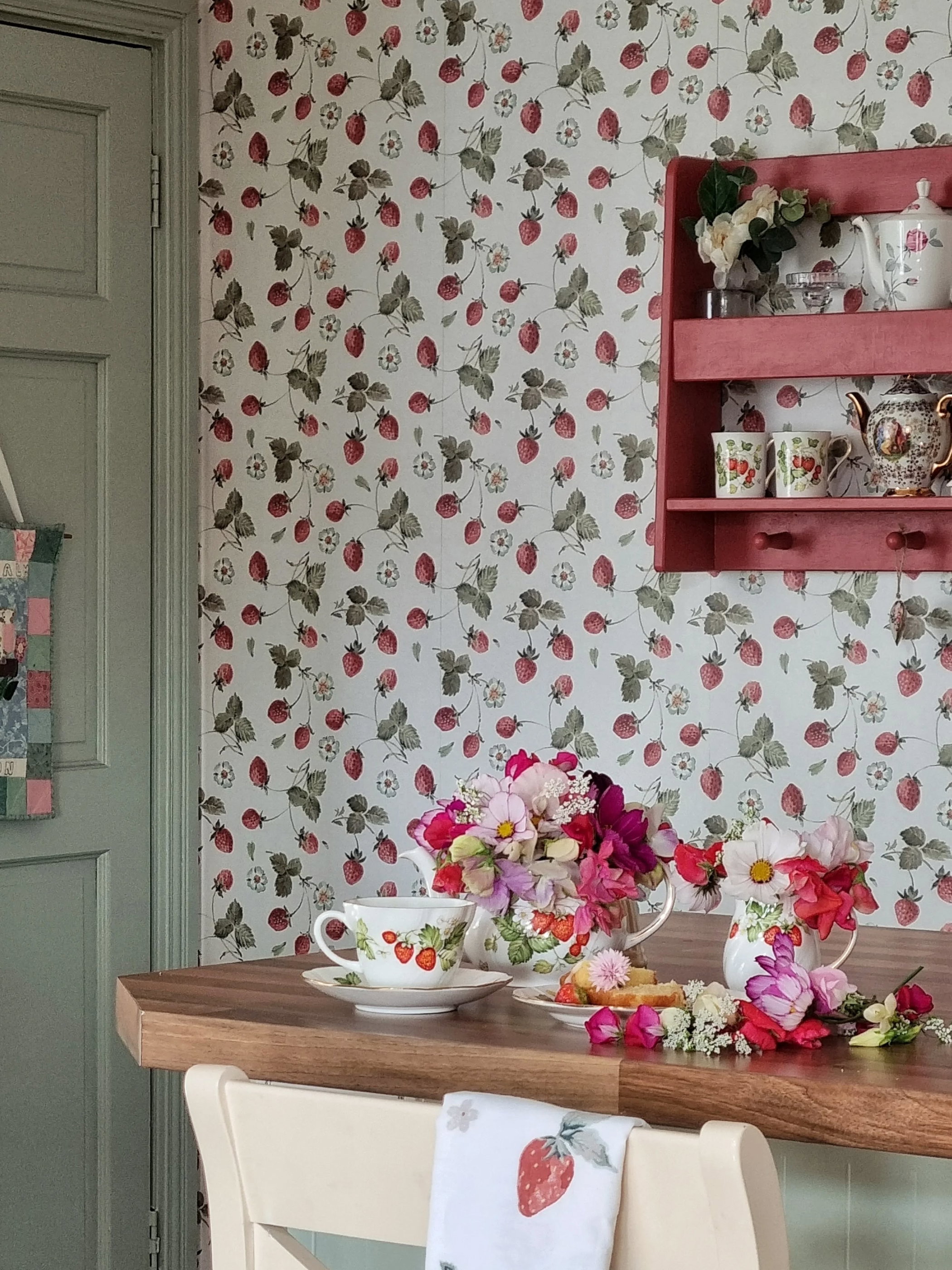 Strawberry-patterned wallpaper adorns a kitchen with teacups, teapots, and flowers on a wooden counter.
