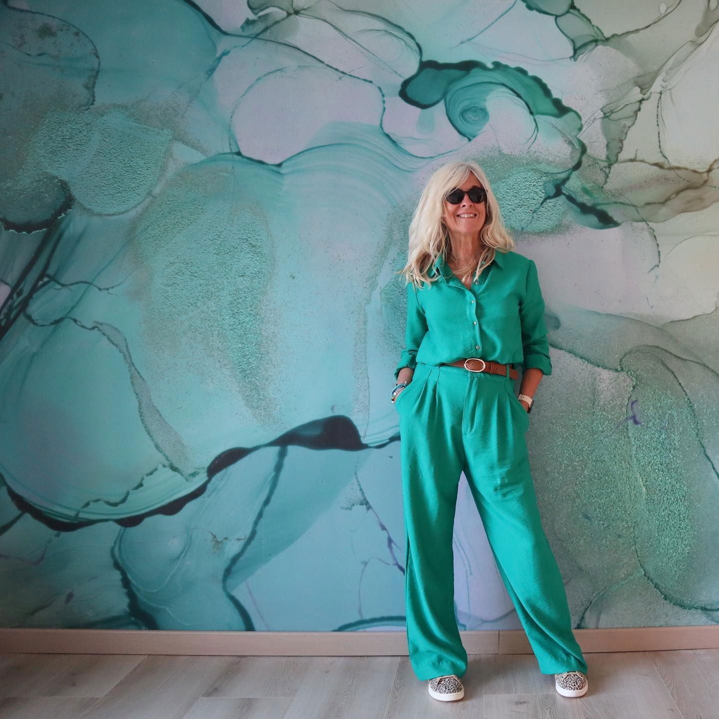 Woman in green linen suit with brown belt and animal print shoes, posing against an abstract teal wall.